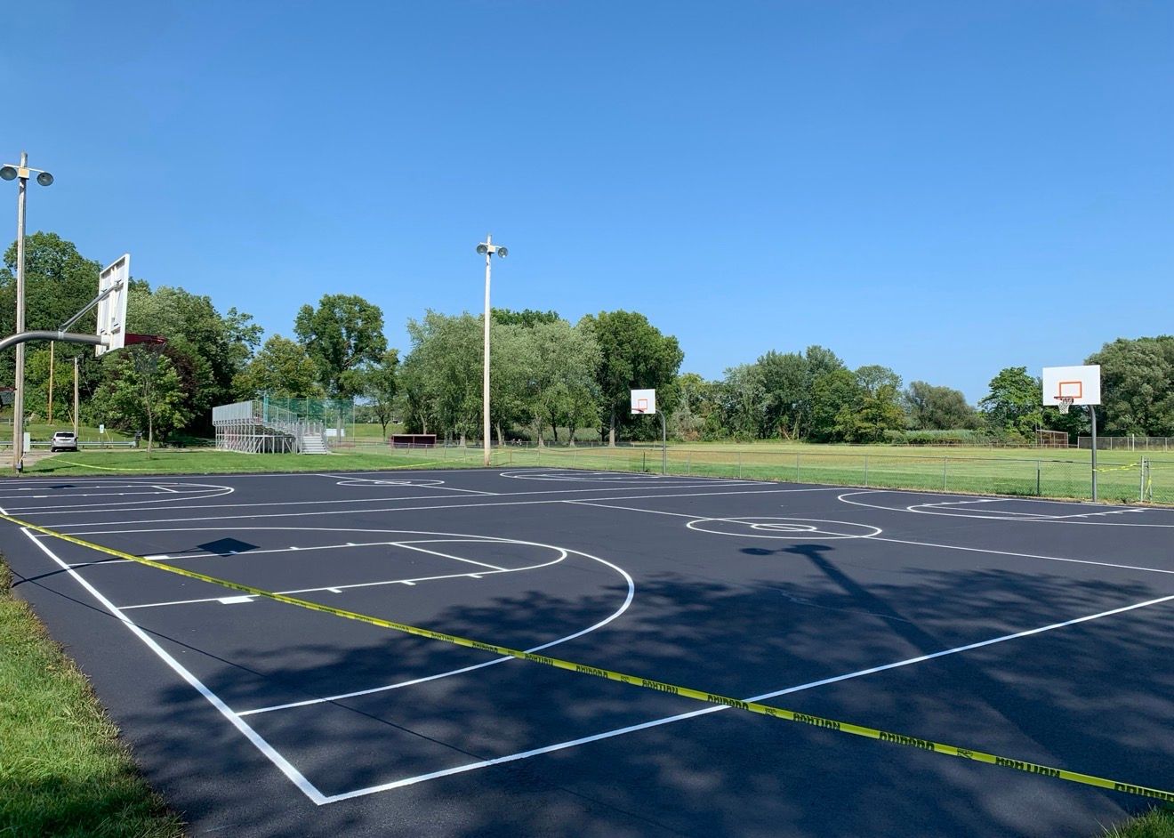 An outdoor basketball court with a freshly paved black surface and white boundary markings under a clear blue sky.