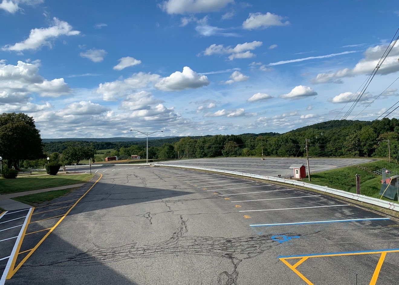 An empty asphalt parking lot with white-lined spaces, marked by yellow curbs under a sunny sky with clouds.