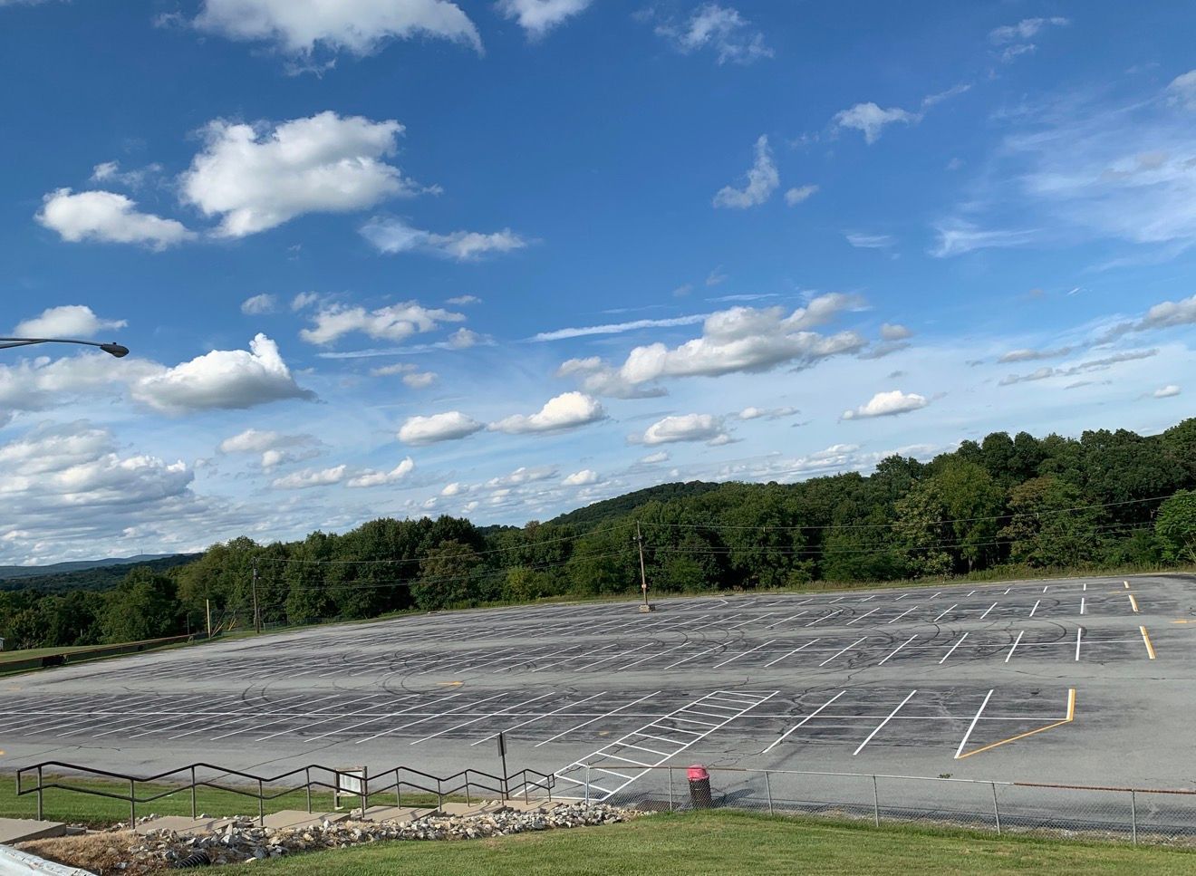 A large, empty paved parking lot with painted lines under a blue sky with scattered clouds, bordered by trees.