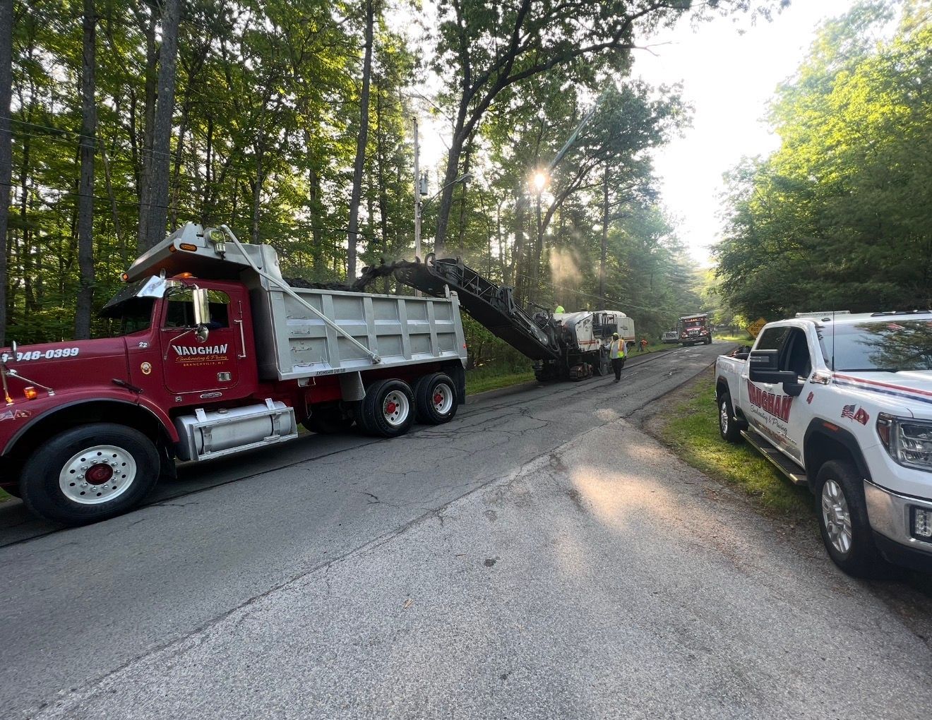 A red dump truck and a white pickup truck on a rural road with a construction machine performing road work.