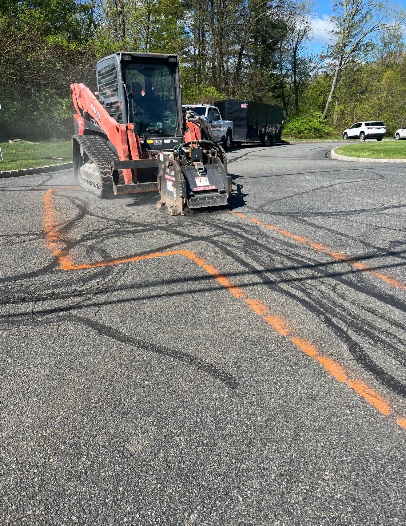 A red skid steer with a concrete saw attachment cuts a line through a paved parking lot marked with orange utility paint.