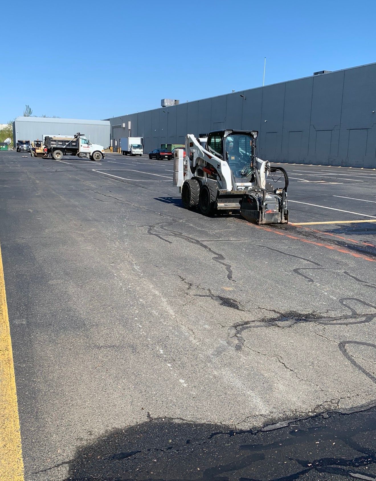 A Bobcat skid-steer with a cold planer attachment works on a large asphalt parking lot near a warehouse building.