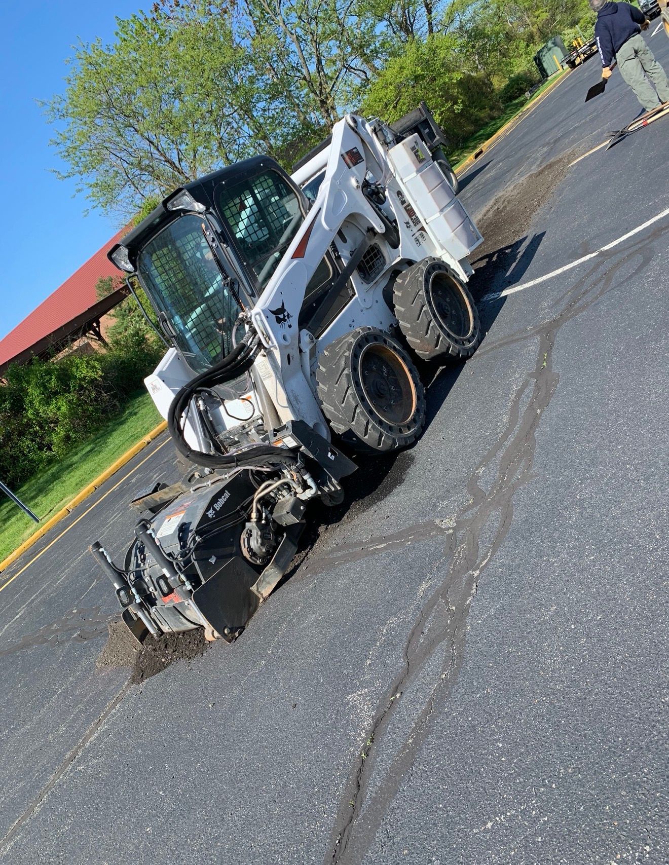 A white Bobcat skid-steer loader with a pavement milling attachment is grinding an asphalt surface in a parking lot.