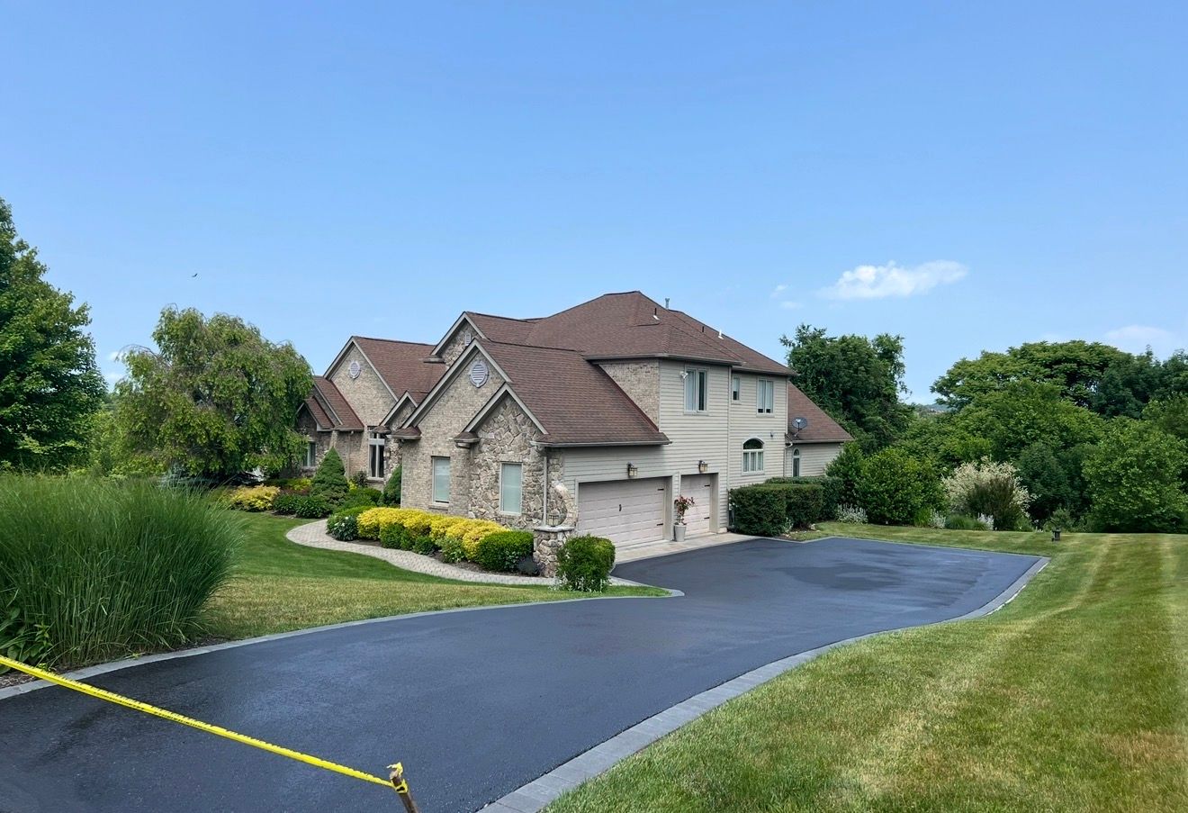 A tan stone and stucco house with a dark roof and paved driveway, surrounded by green lawns and trees under a blue sky.