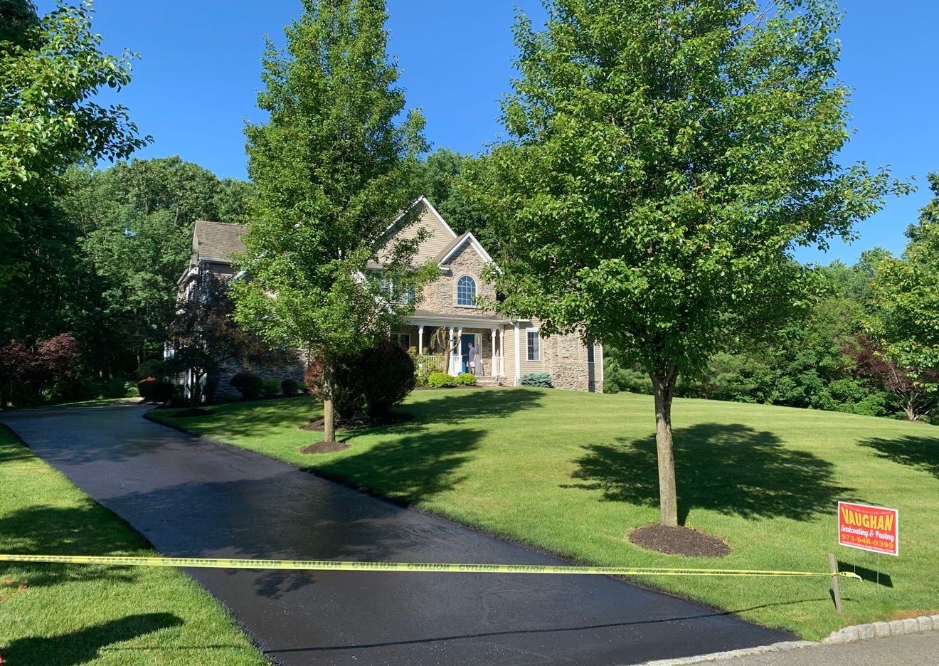 A large house with a freshly paved asphalt driveway, surrounded by green lawns and trees under a clear blue sky.