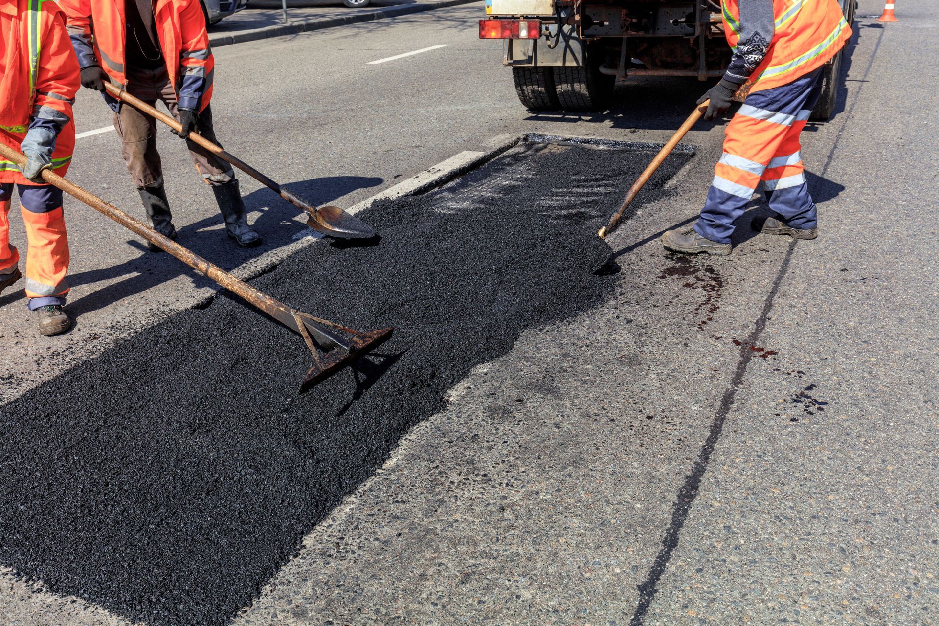 Road workers in high-visibility uniforms use rakes to spread fresh asphalt during a road repair.