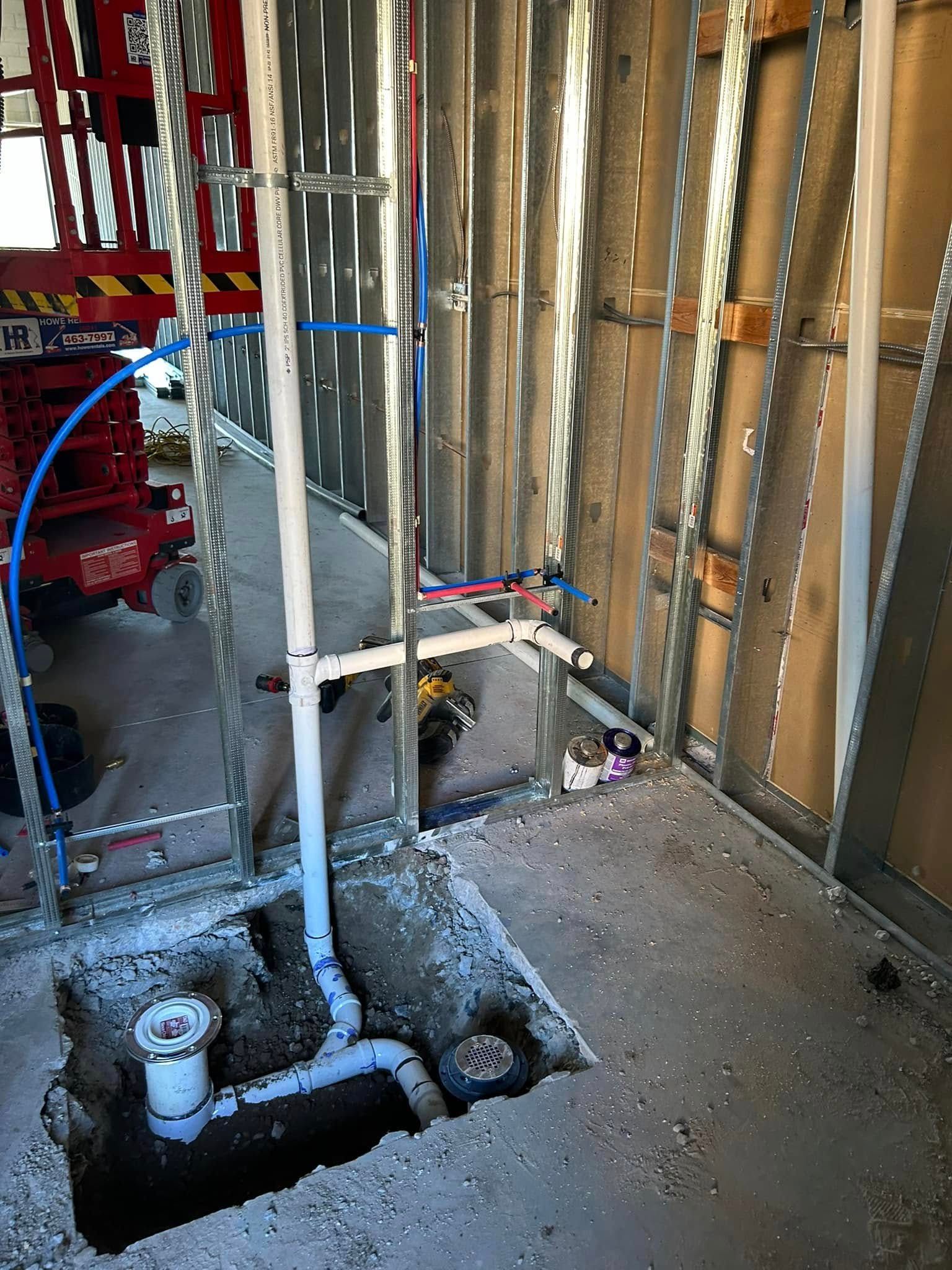 Construction site with exposed plumbing and wall studs. A hole in the concrete floor reveals pipes and drains.