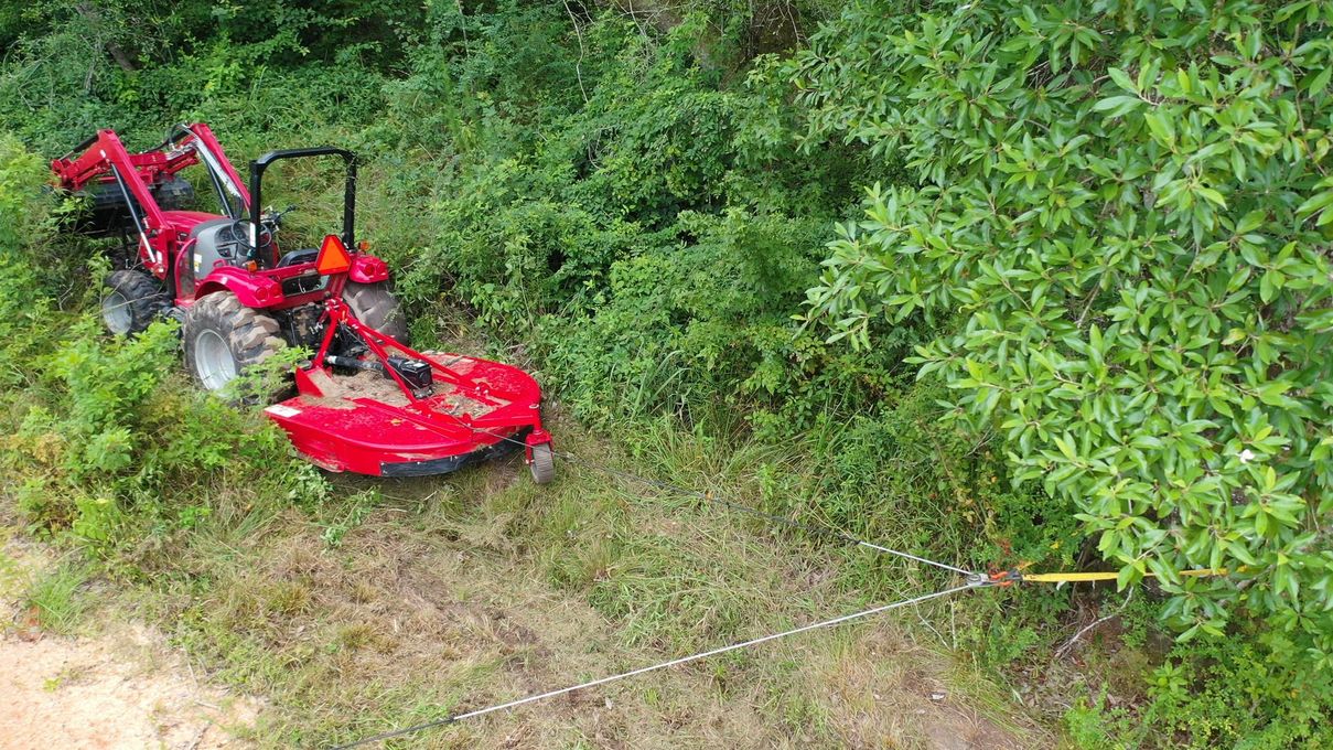 Red tractor overturned in dense green brush, partially on a dirt path.