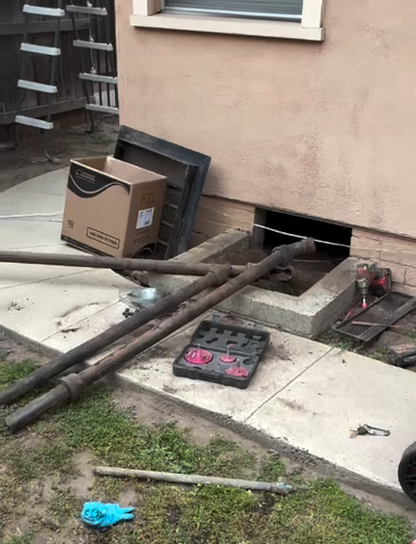 Rusted metal pipes run into a basement wall opening beside a box and tools on a concrete patio.