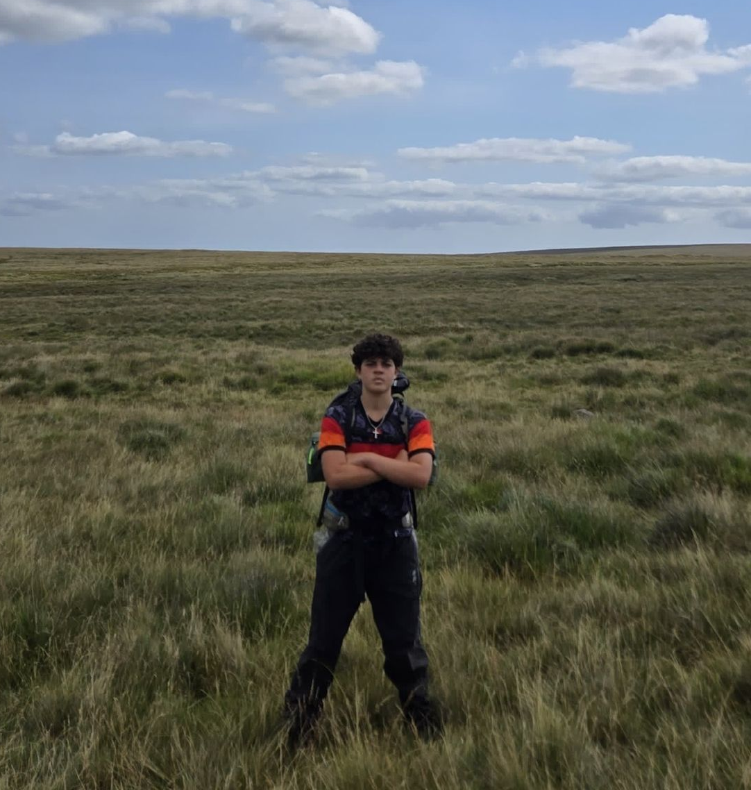 16 year old Christopher stands in a wide grassy landscape wearing hiking gear and a backpack, preparing for his challenge to climb Mount Kilimanjaro and raise funds for The Sparkle Foundation's programmes in Malawi.