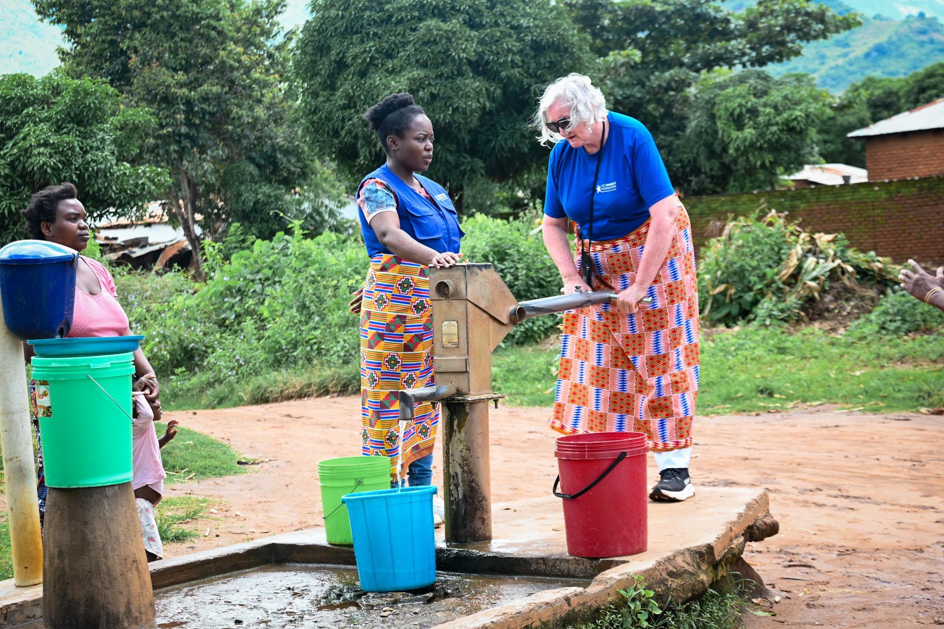 Leith stands with Frances at a rural community borehole. Leith is actively using the hand pump to draw water while Frances observes beside her during a community visit in Zomba, Malawi.