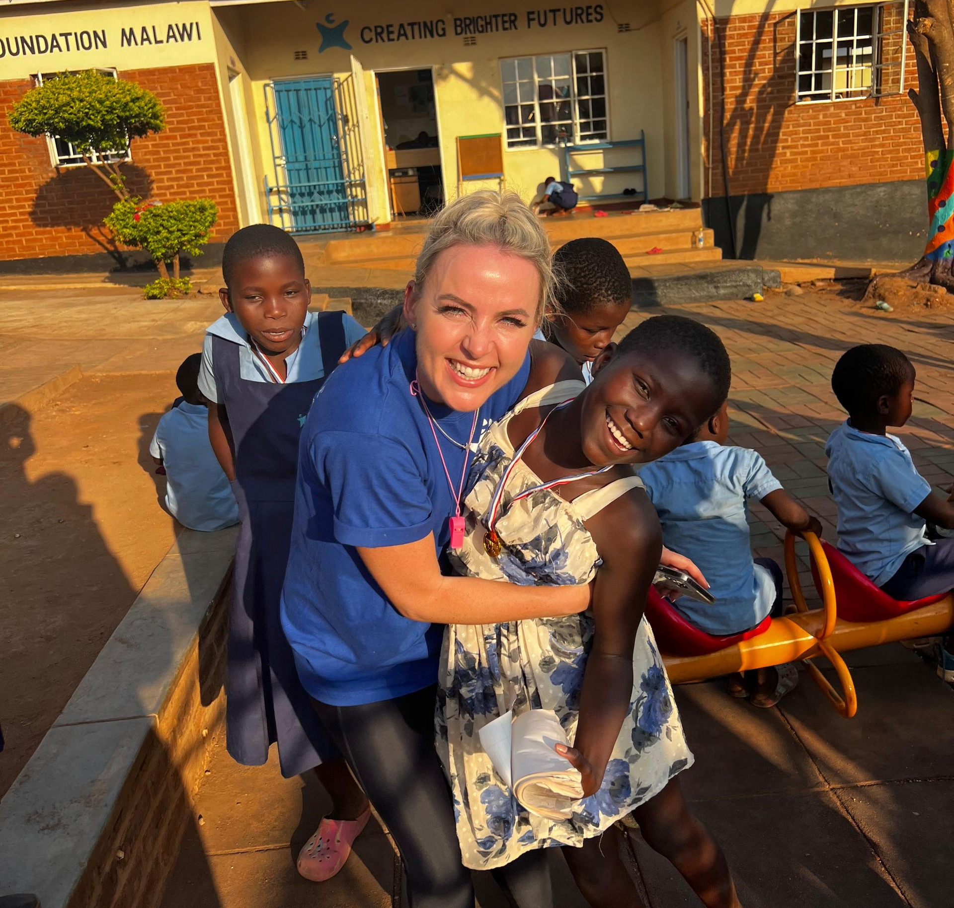 Kelly Lundberg hugs a smiling child outside The Sparkle Foundation's education centre in Malawi during her volunteer visit, surrounded by children playing in the school courtyard.