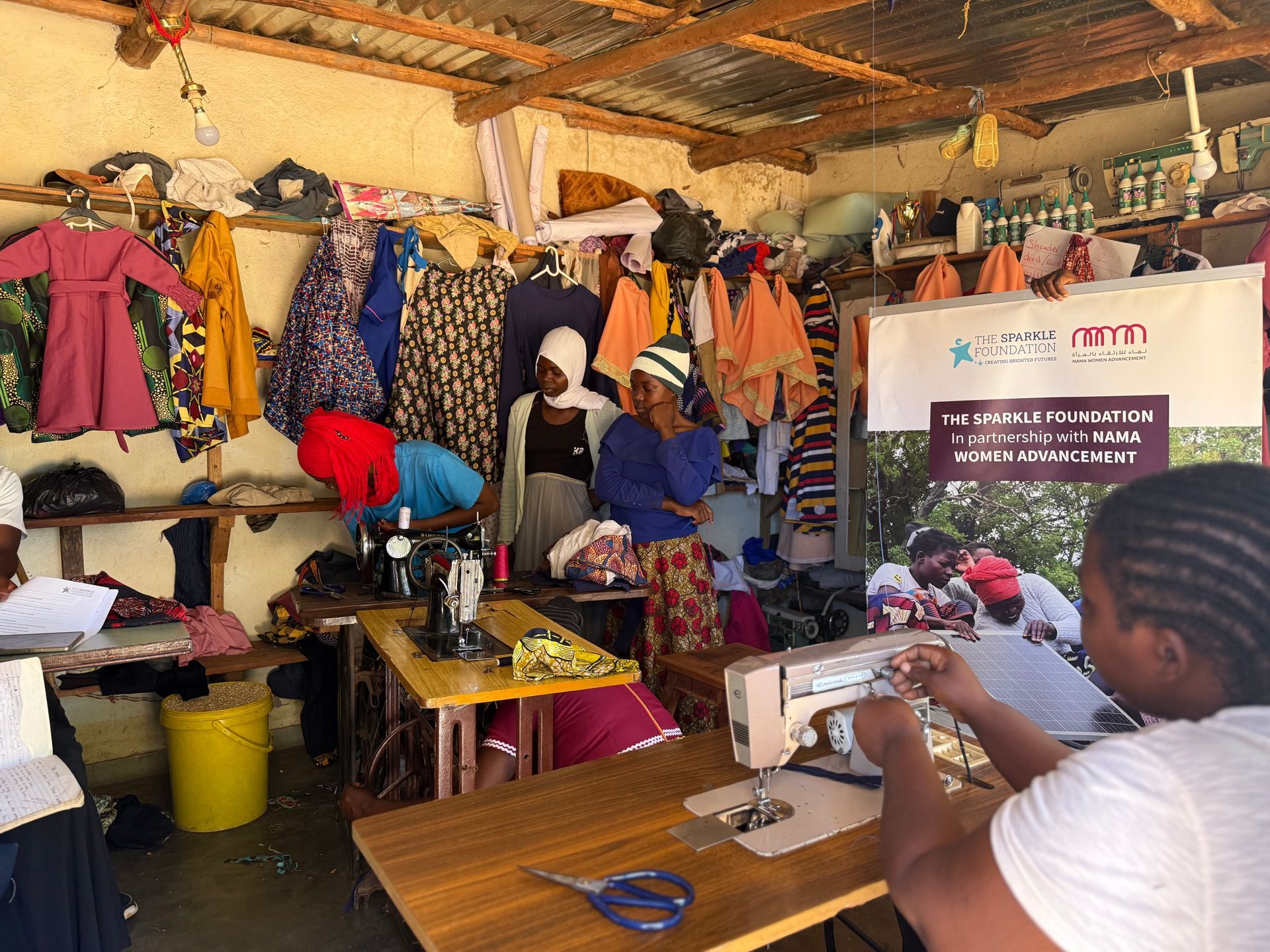 Participants gather inside the Spark to Success tailoring training centre in Sakata, Zomba District, as trainees demonstrate sewing skills and discuss garment production. Dresses, skirts, and other handmade items are displayed throughout the workshop alongside a banner representing the partnership between The Sparkle Foundation and NAMA Women Advancement.