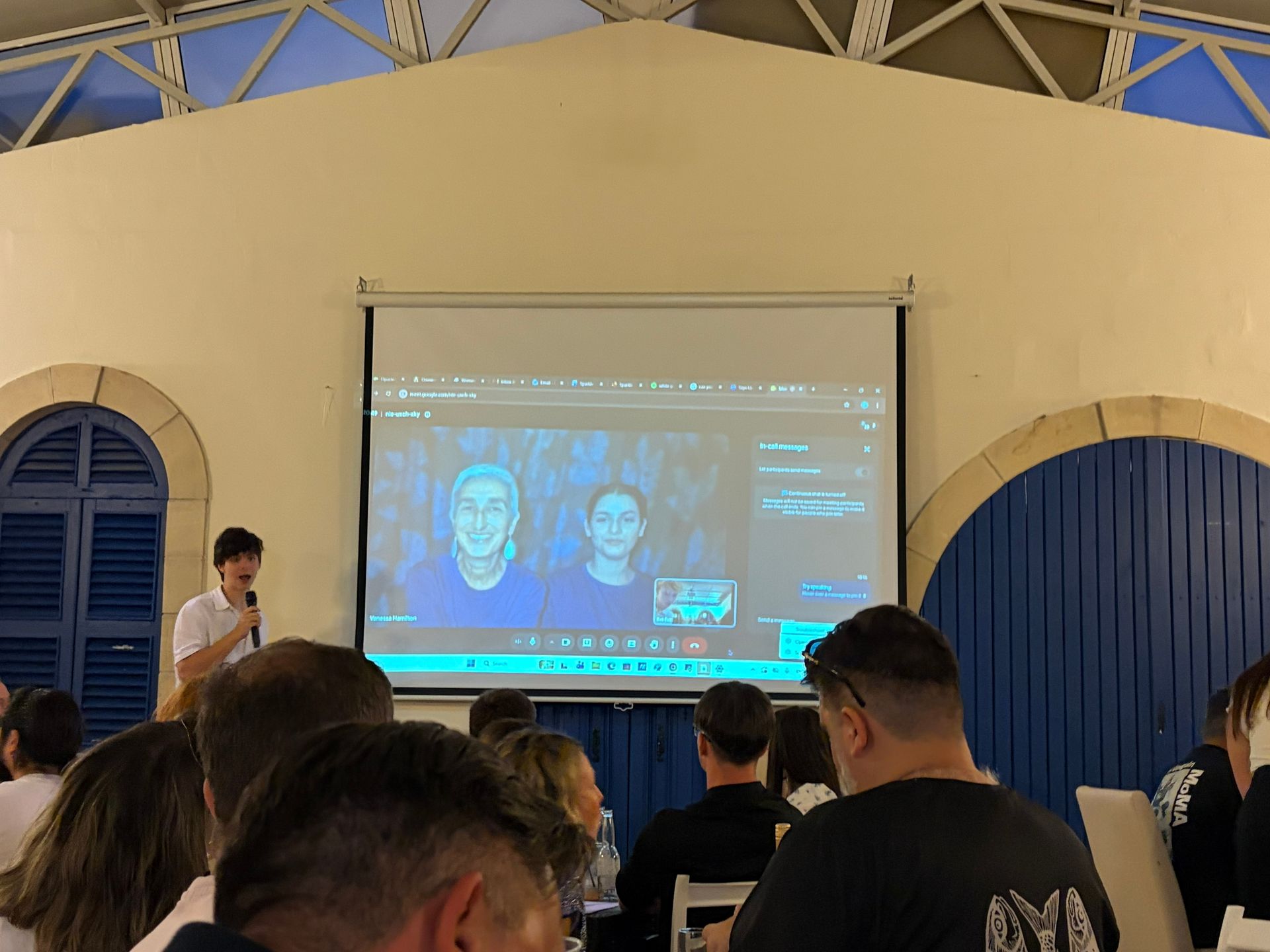 A student speaks into a microphone while a large projector screen displays a live video call with The Sparkle Foundation's Project Coordinator, Vanessa Hamilton, and a colleague in Malawi. Audience members are seated at tables during a community fundraising quiz night, watching the presentation.