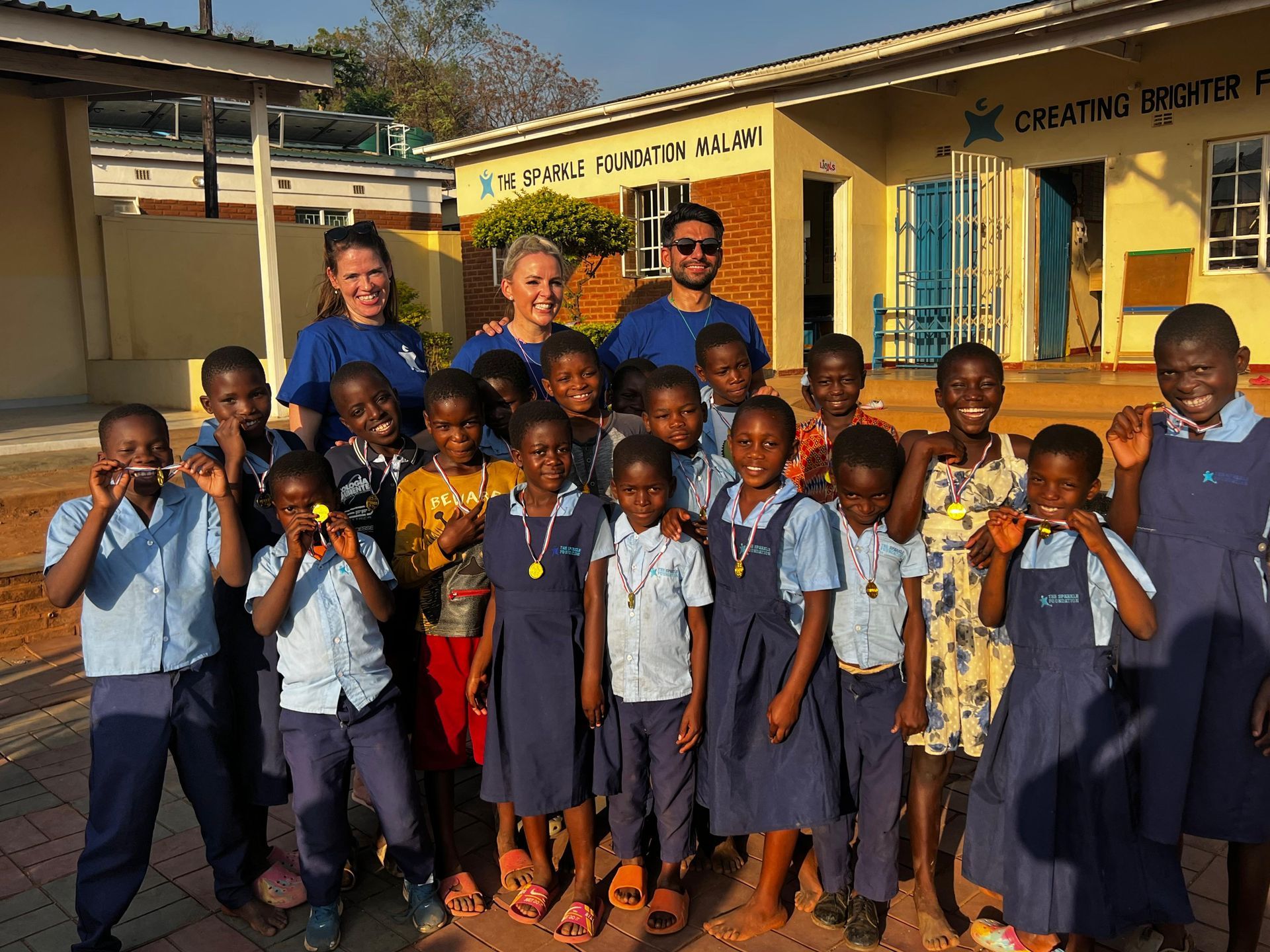 Kelly Lundberg and fellow volunteers pose with smiling children wearing medals outside The Sparkle Foundation's education centre in Malawi after a community sports day.