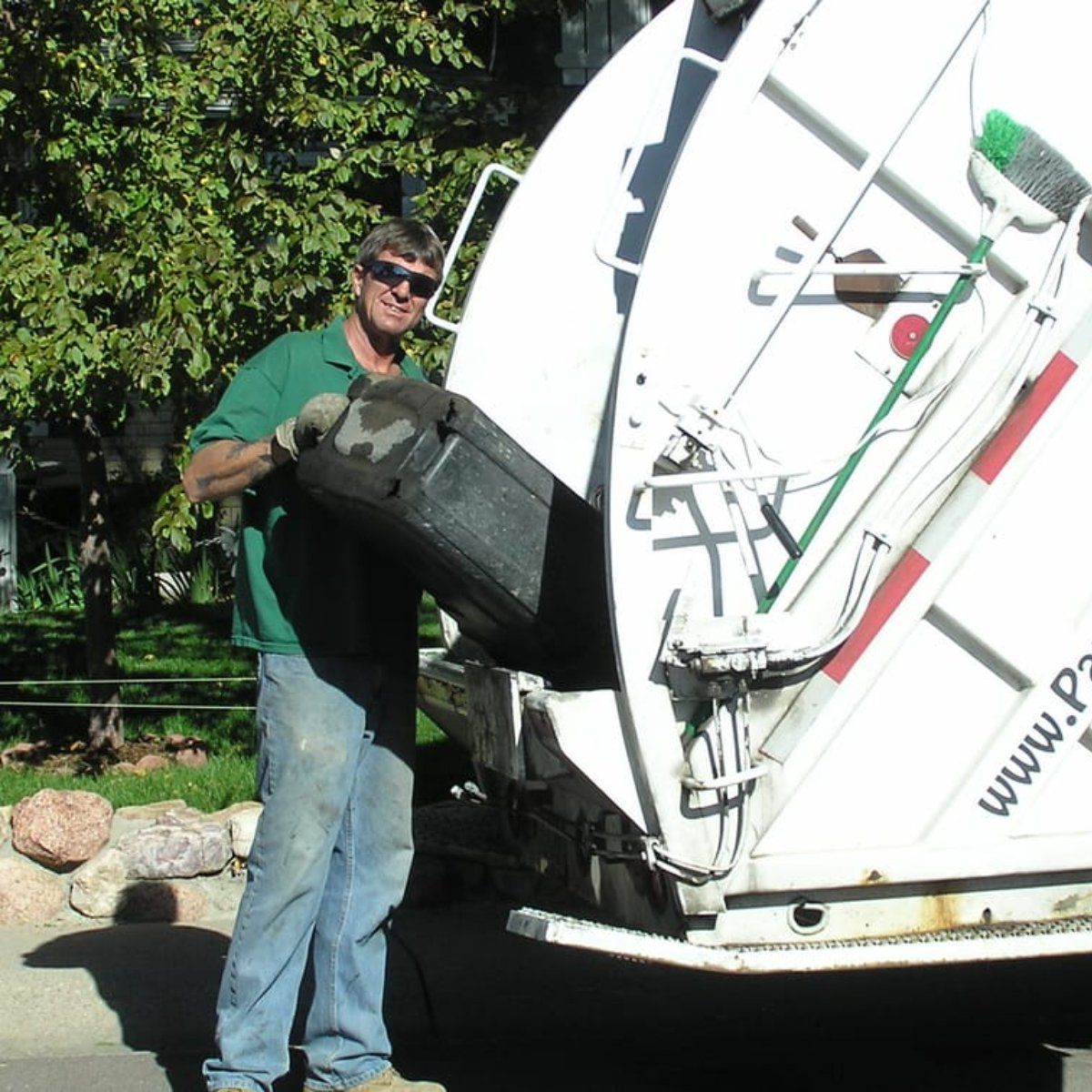 A man in a green shirt is loading a trash can into a garbage truck