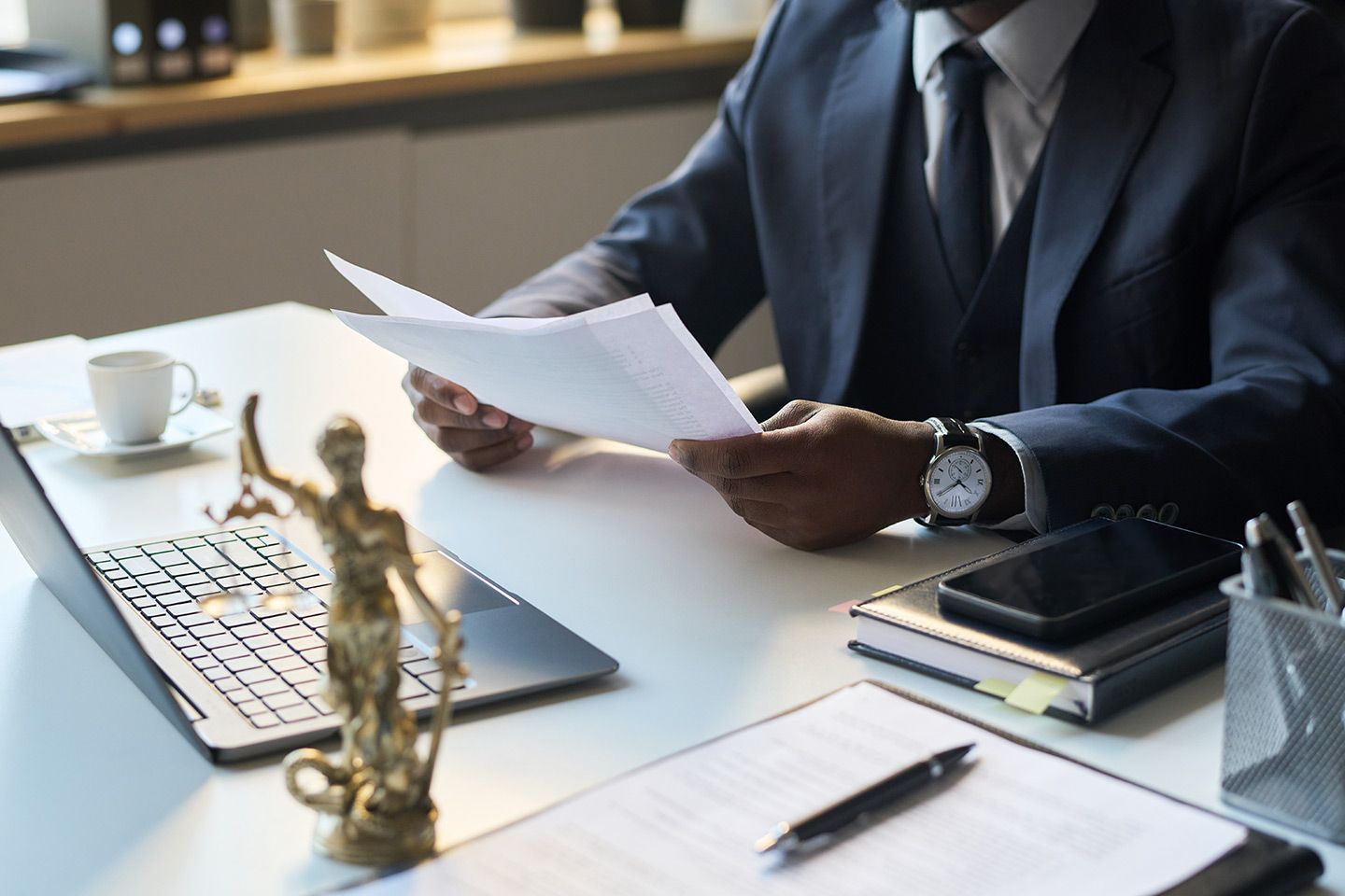 Person in suit reviewing documents at a desk, scales of justice statue, laptop, phone, and coffee cup.