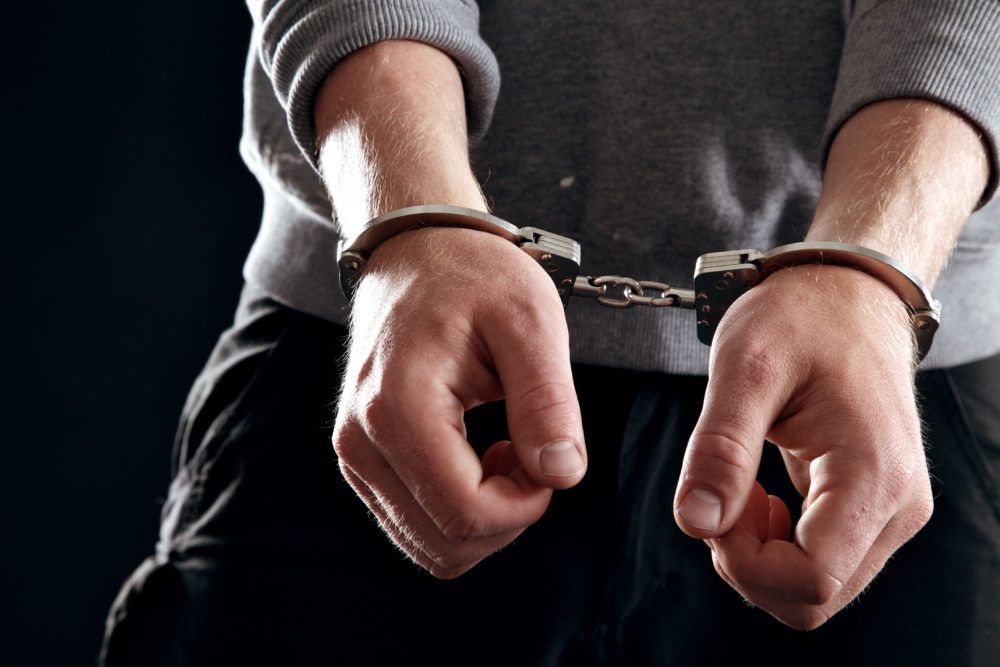 A person in a gray sweatshirt with their hands cuffed together in front of them against a dark background.