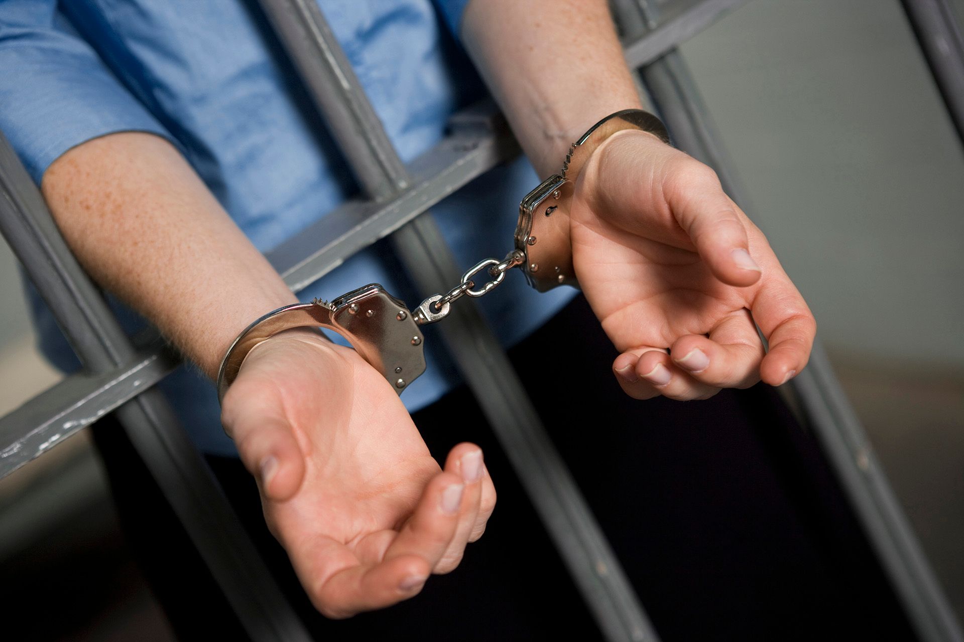 A person in a blue shirt wearing handcuffs, positioned behind gray metal prison bars.