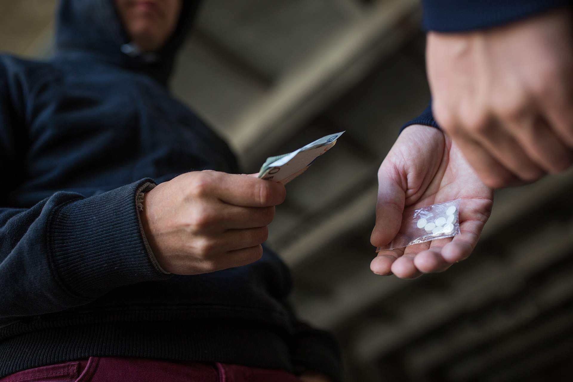 Two people exchange a banknote and small pills in a dim outdoor setting