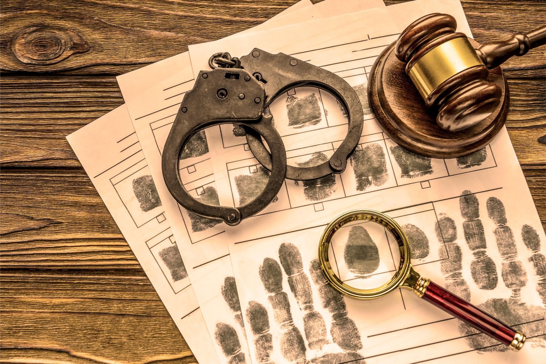 Handcuffs, magnifying glass, gavel on a wooden surface atop fingerprint documents, signifying crime scene investigation.