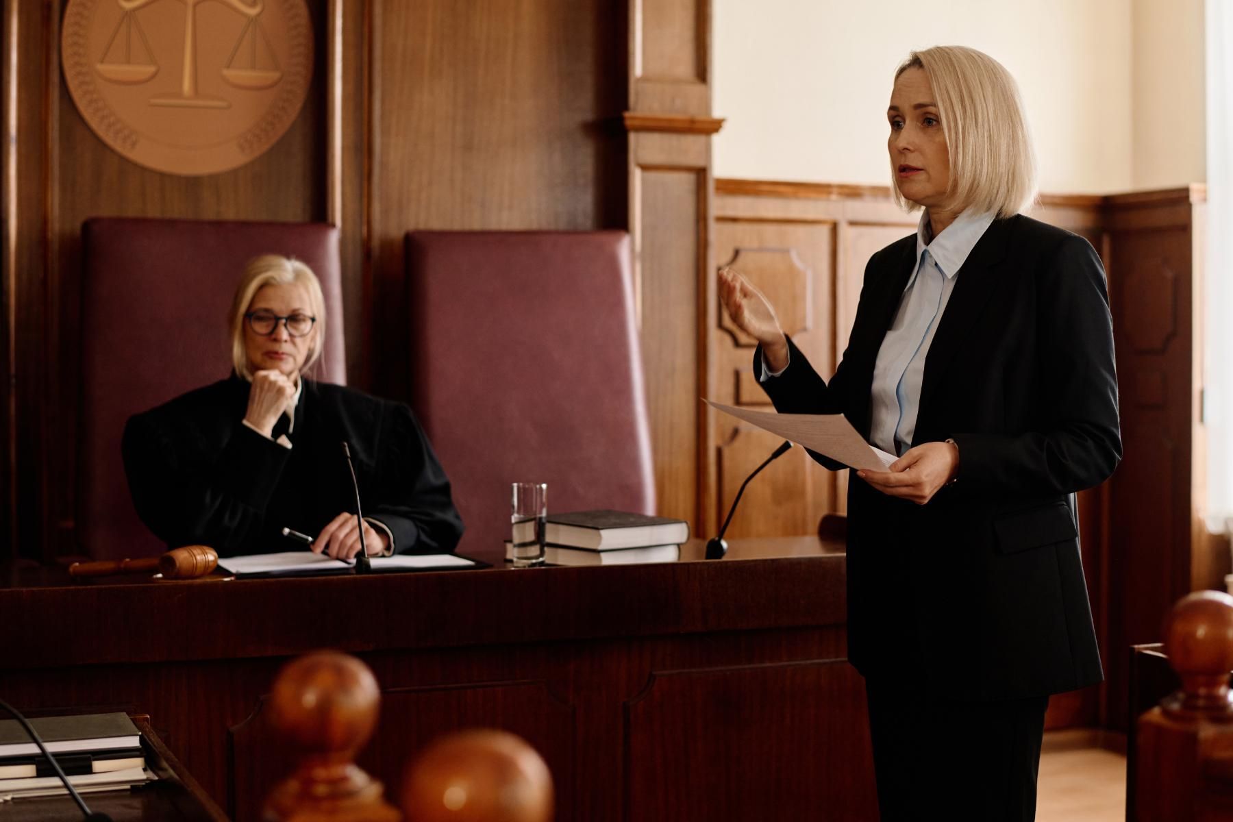 A lawyer in a dark suit speaks in a courtroom. A judge in black robes listens.