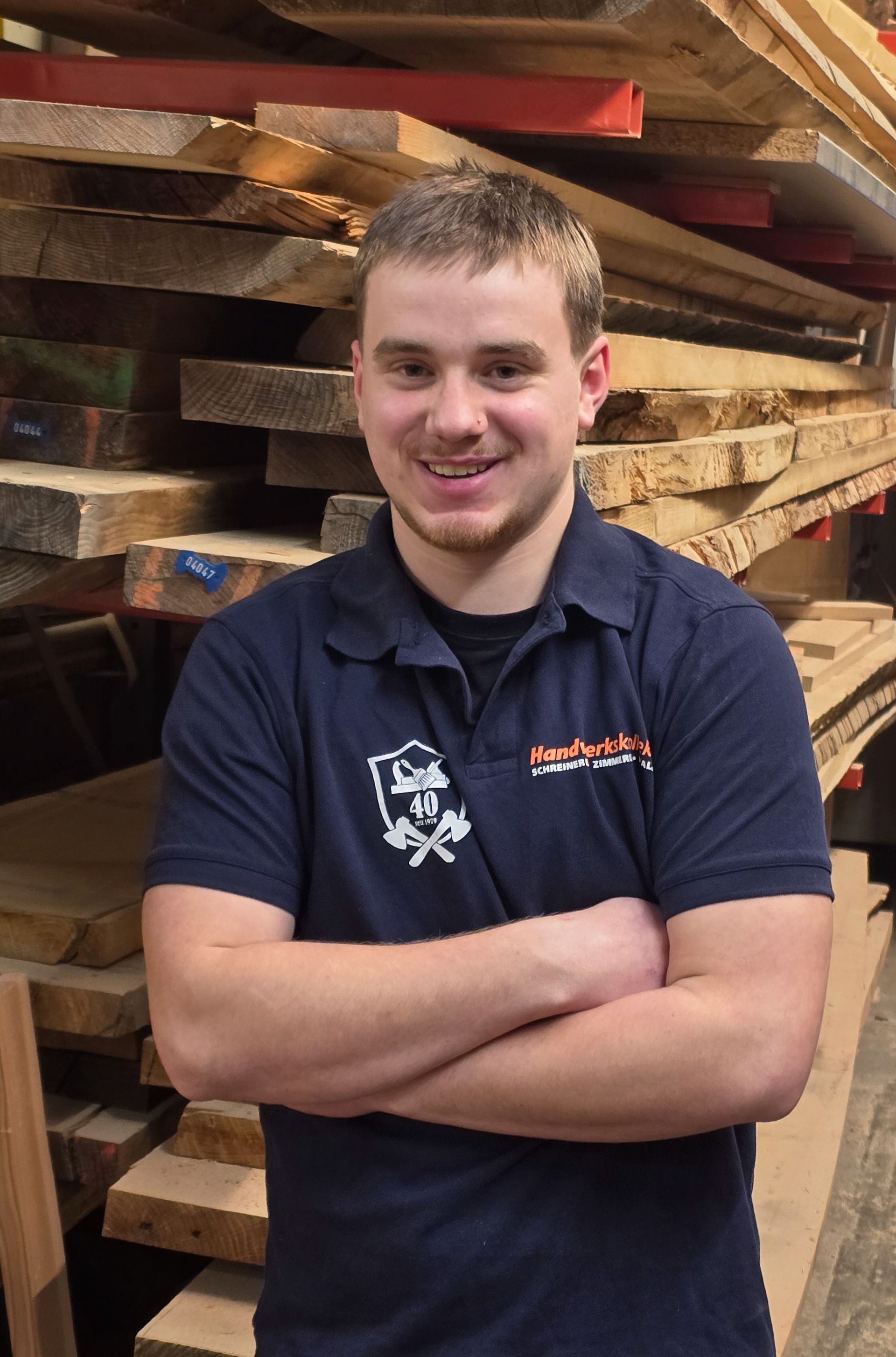 Ein Mann mit verschränkten Armen lächelt vor einem Stapel Holz. Er trägt ein dunkelblaues Hemd mit einem Logo.