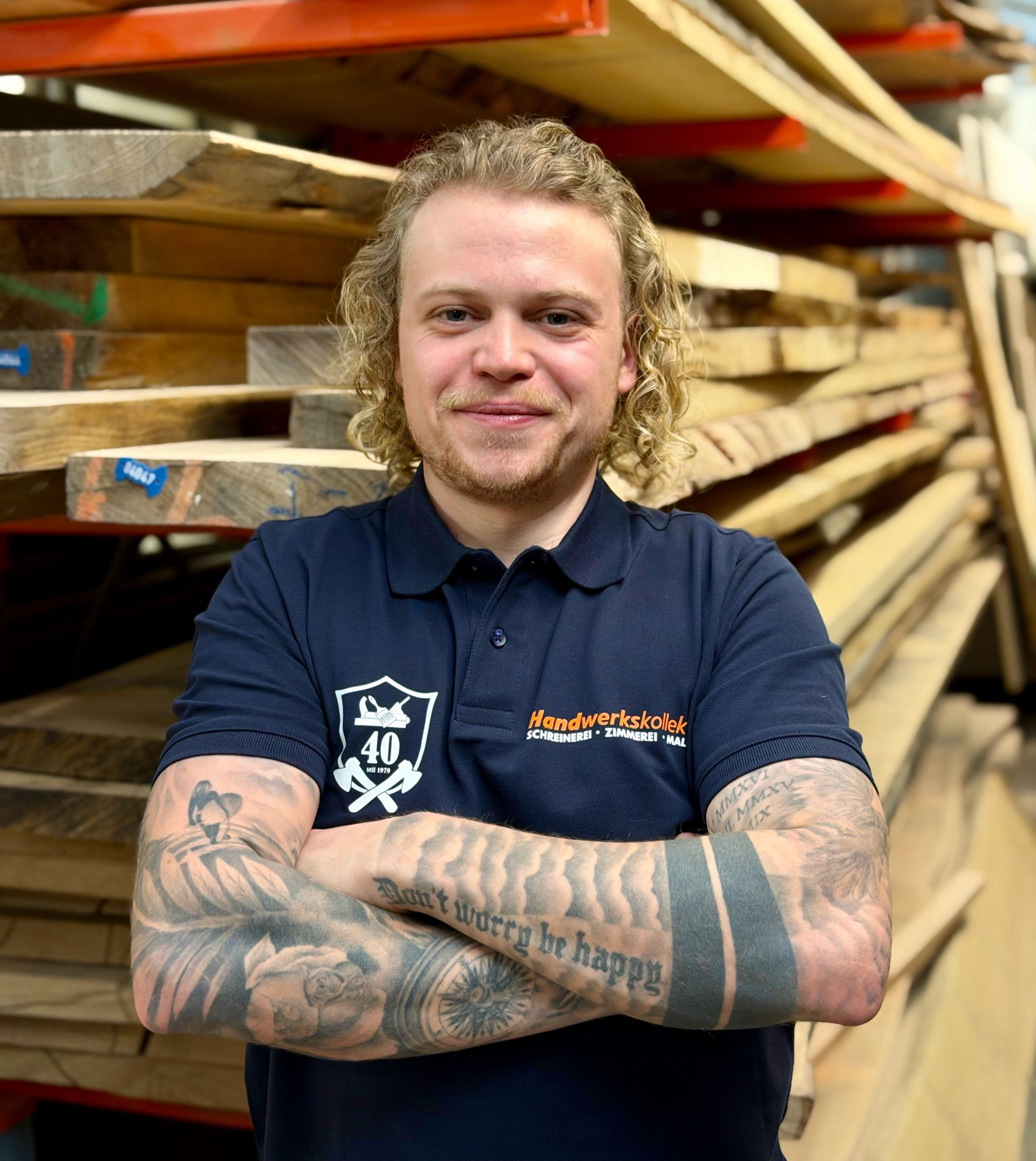 Ein Mann mit Armtätowierungen und verschränkten Armen lächelt in einem Sägewerk und trägt ein Poloshirt mit Logo.