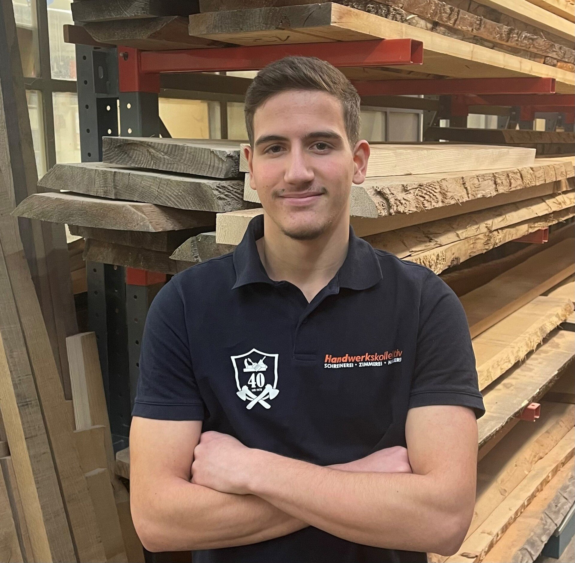 Ein junger Mann mit verschränkten Armen steht vor einem Stapel Holz