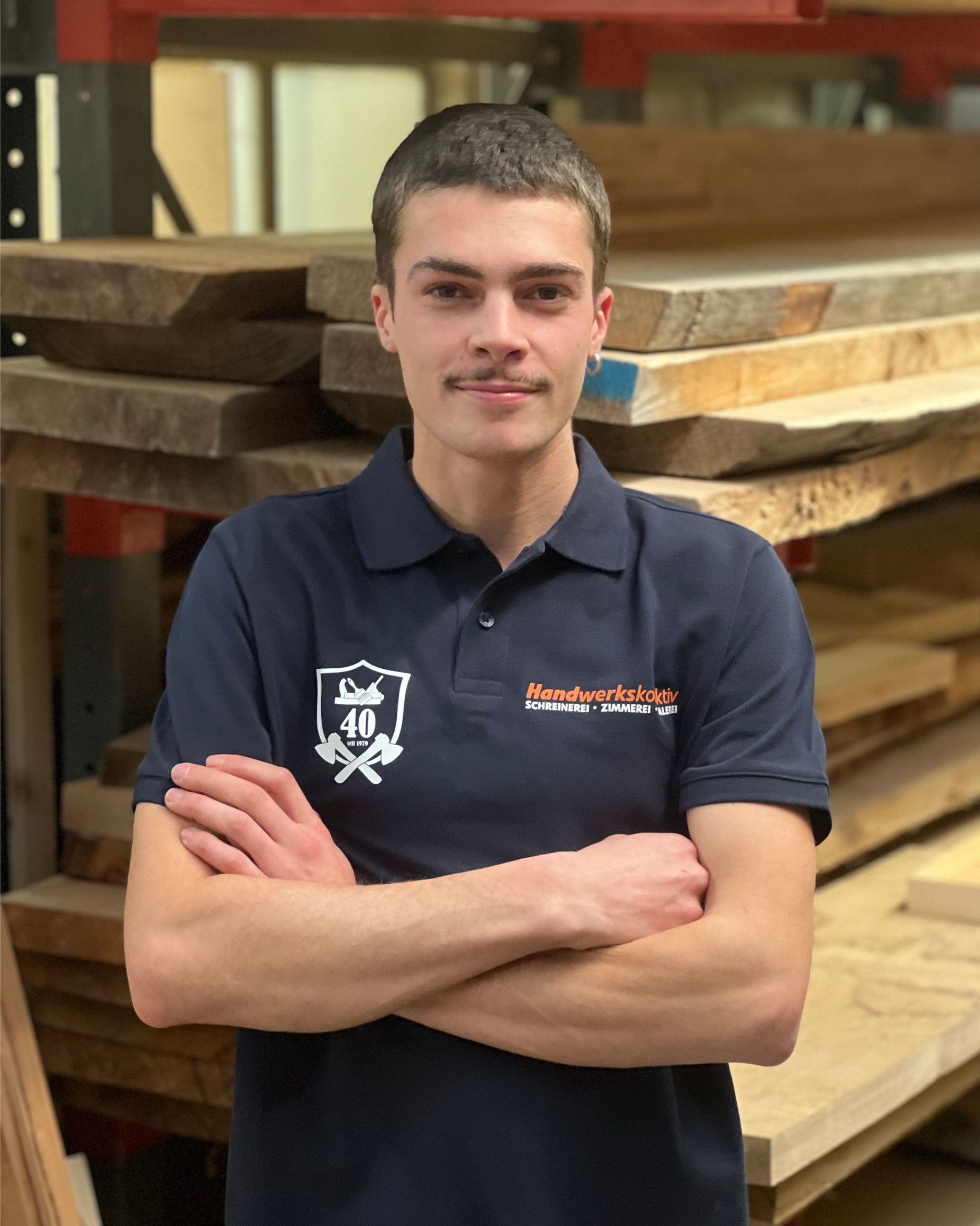 Ein junger Mann mit Schnauz steht vor einem Holzstapel.