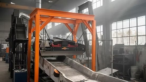 Máquina industrial naranja sobre una cinta transportadora en un taller de fábrica, con una carretilla elevadora al lado.