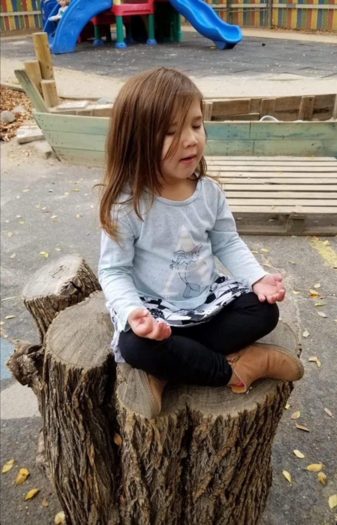 A little girl is sitting on a tree stump in a lotus position.