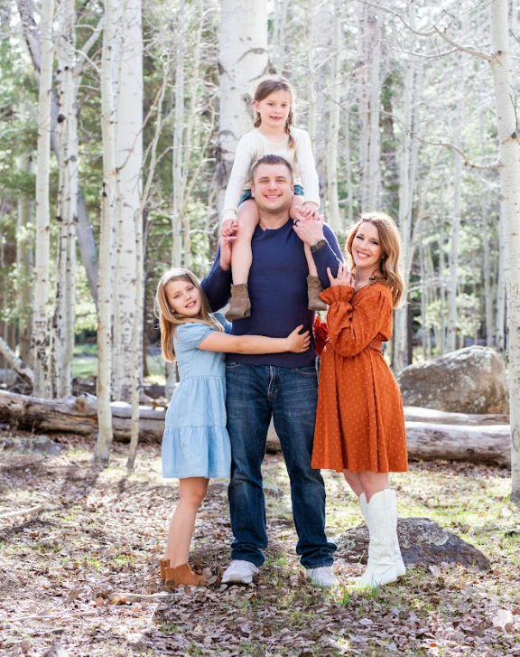 Family posing in a forest. Man with girl on shoulders, two girls, and woman in orange dress. Aspen trees in background.