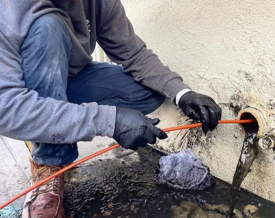 Worker in gray clothes and black gloves clearing a clogged pipe with an orange hose beside a wall.