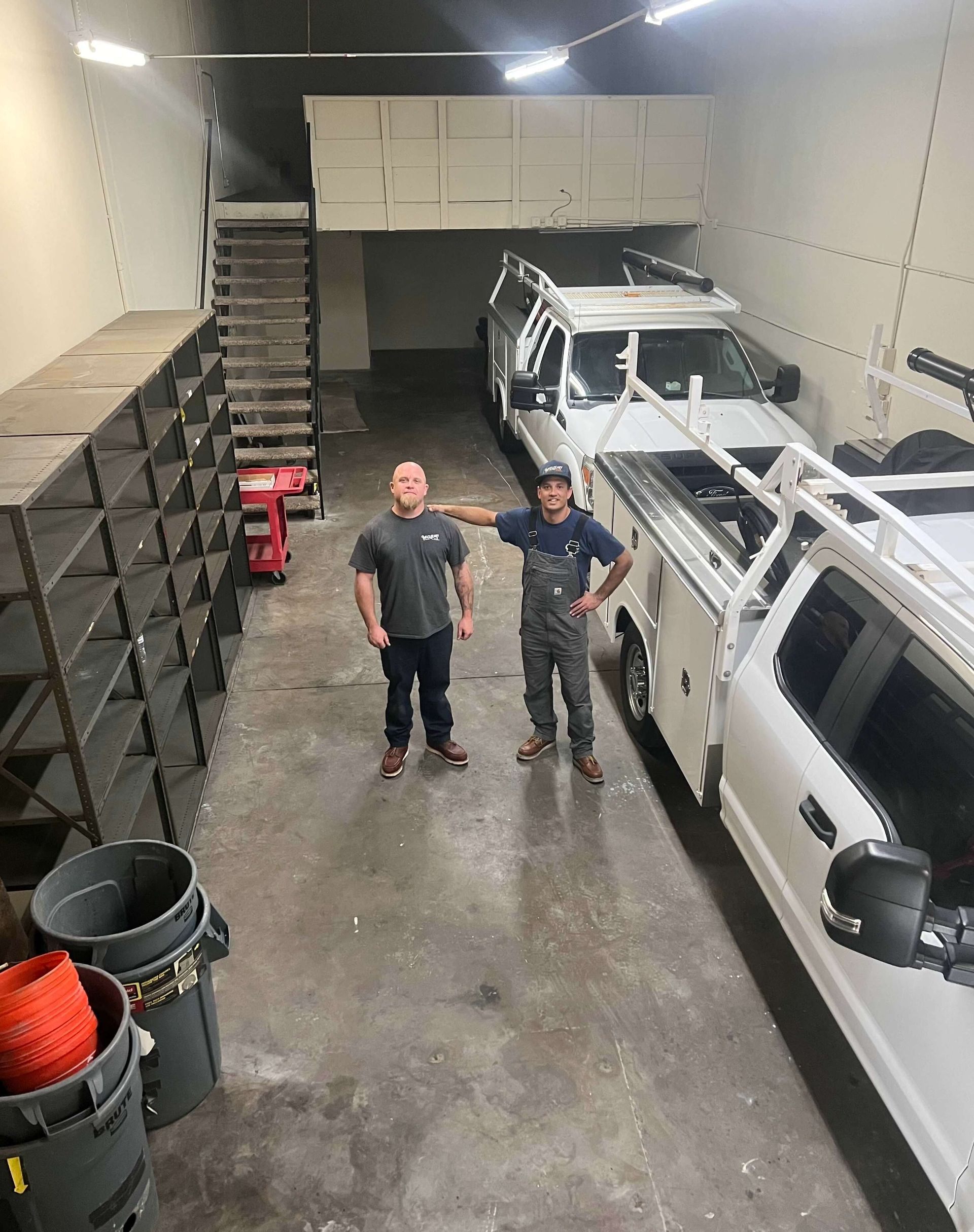 Two men standing in a garage beside a white utility truck and stacked storage bins.