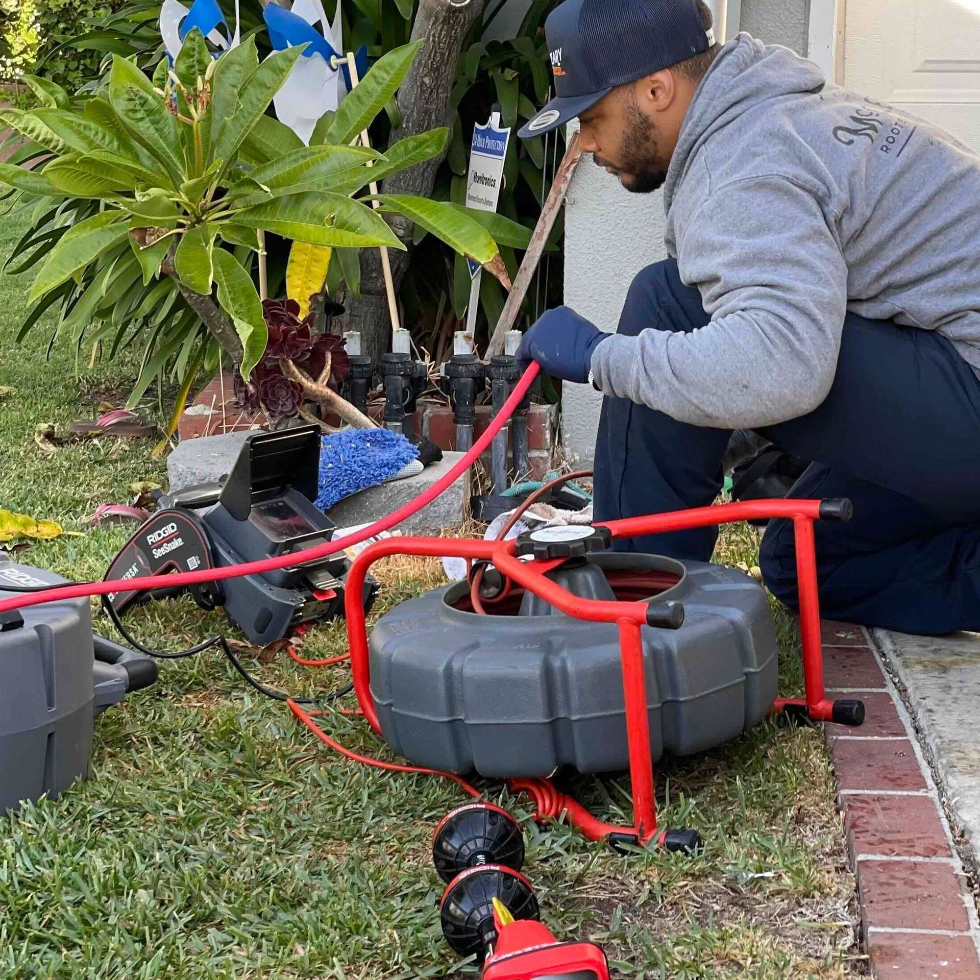 Worker in gray hoodie kneels beside red plumbing tools on grass near a house foundation.