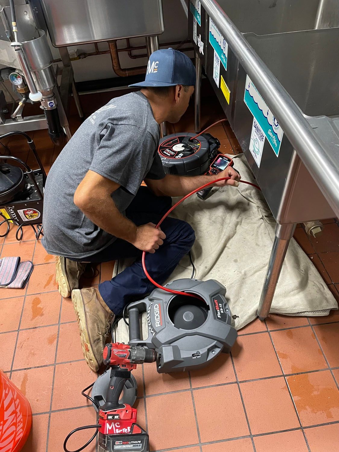 Worker kneels on tiled kitchen floor, using a drain cleaning machine near stainless steel equipment.