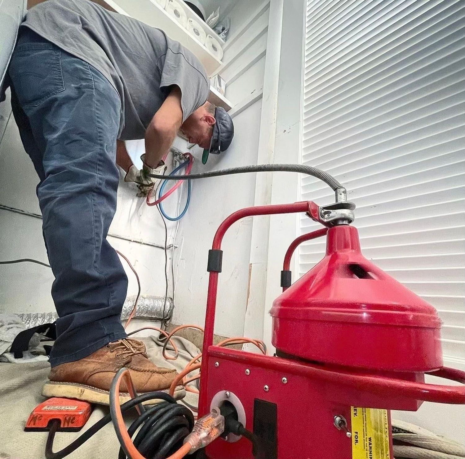 Worker using a red vacuum on a floor, bent over near a wall in a utility area.