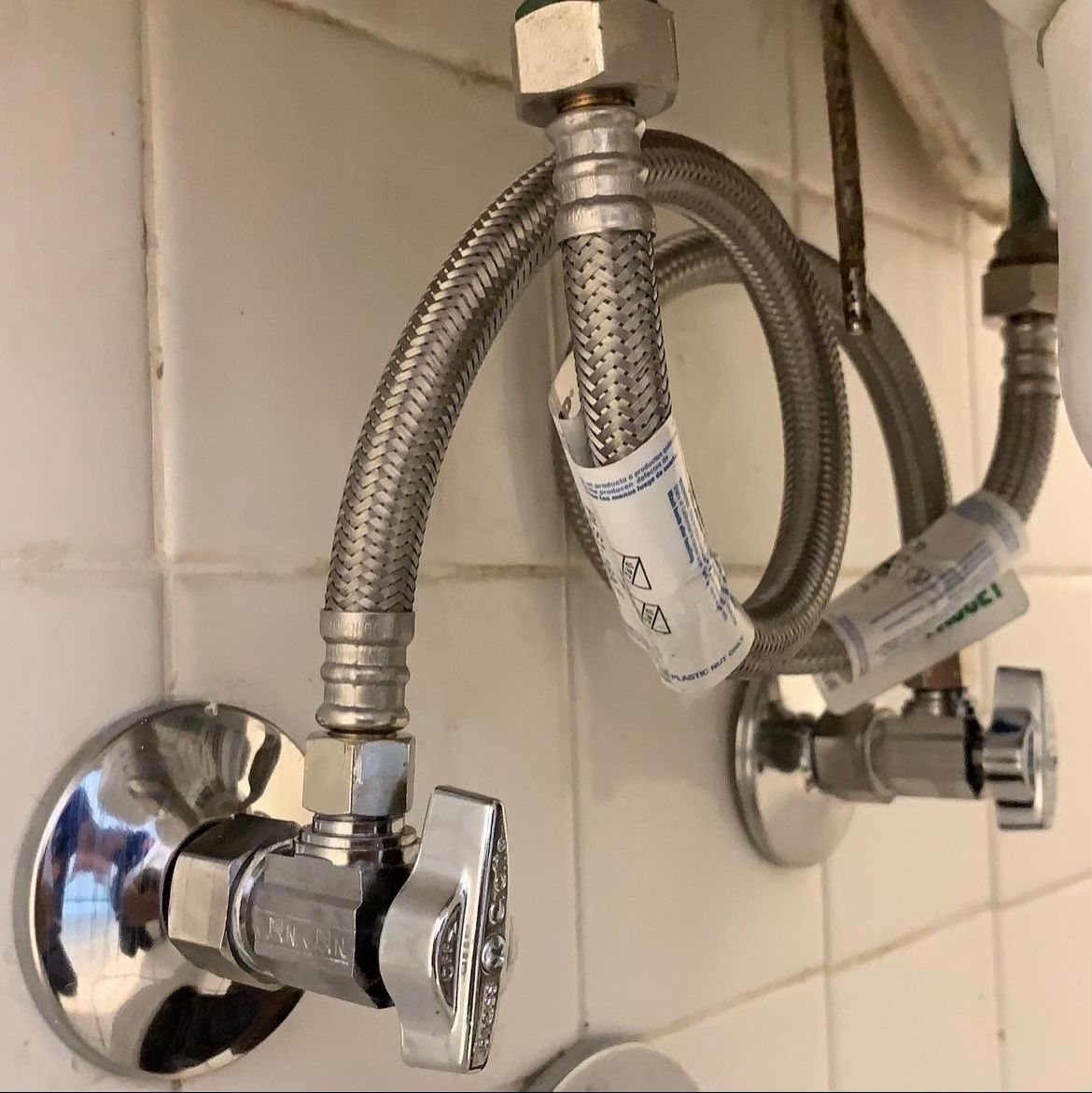 Chrome wall-mounted shower valves with flexible metal supply hoses against white tile.