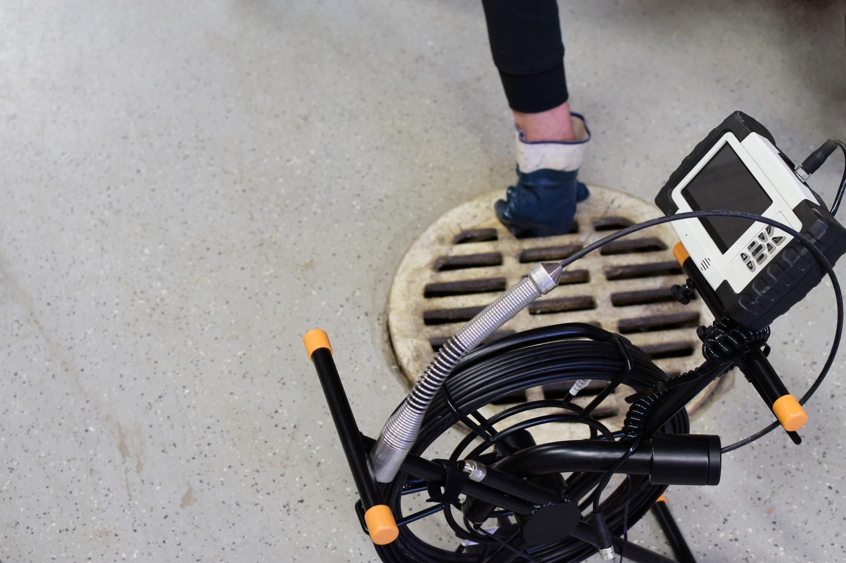 Person standing beside a storm drain with a black cable reel and equipment cart on a concrete surface