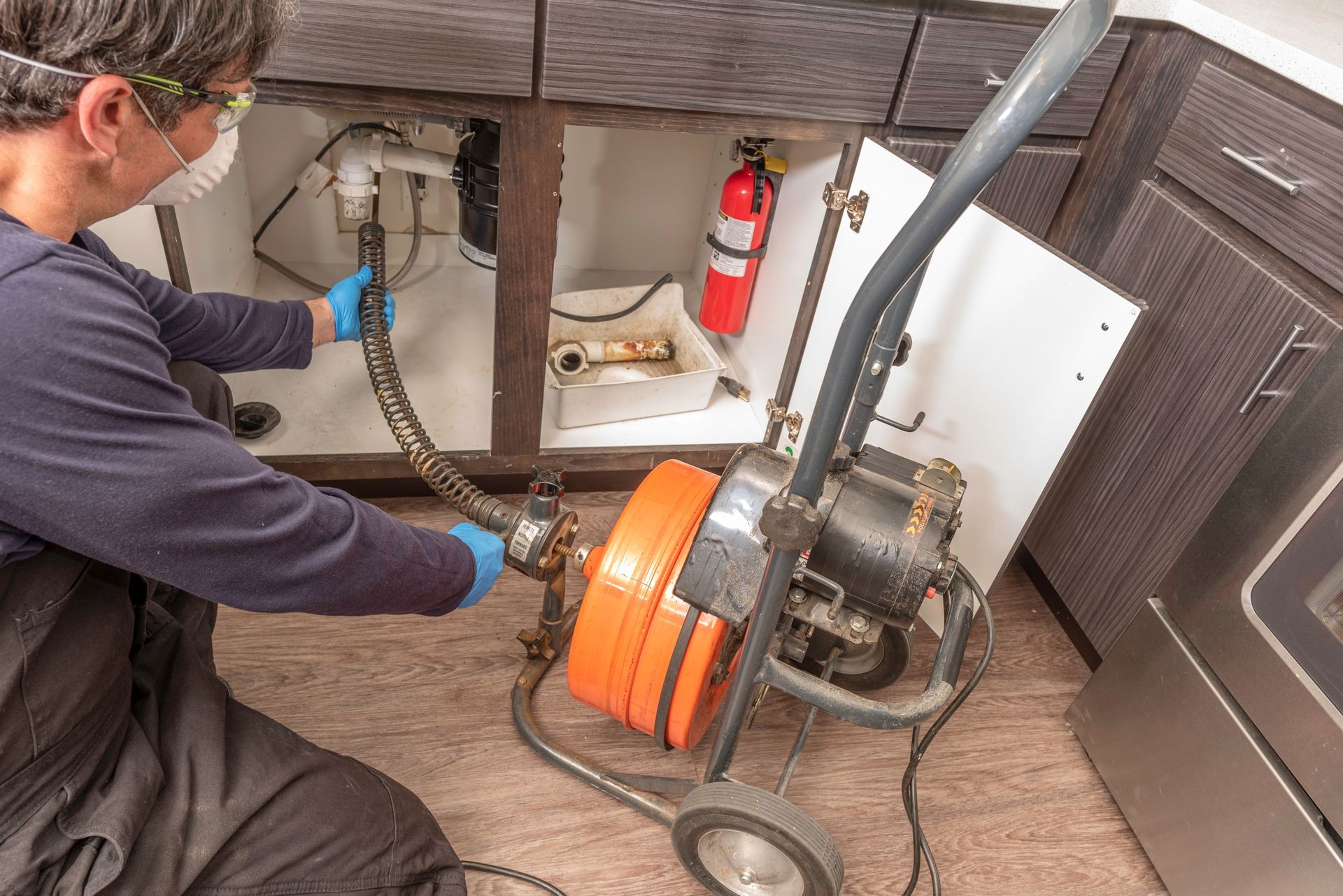 Worker unclogging a floor drain with a power auger beside a fire extinguisher.