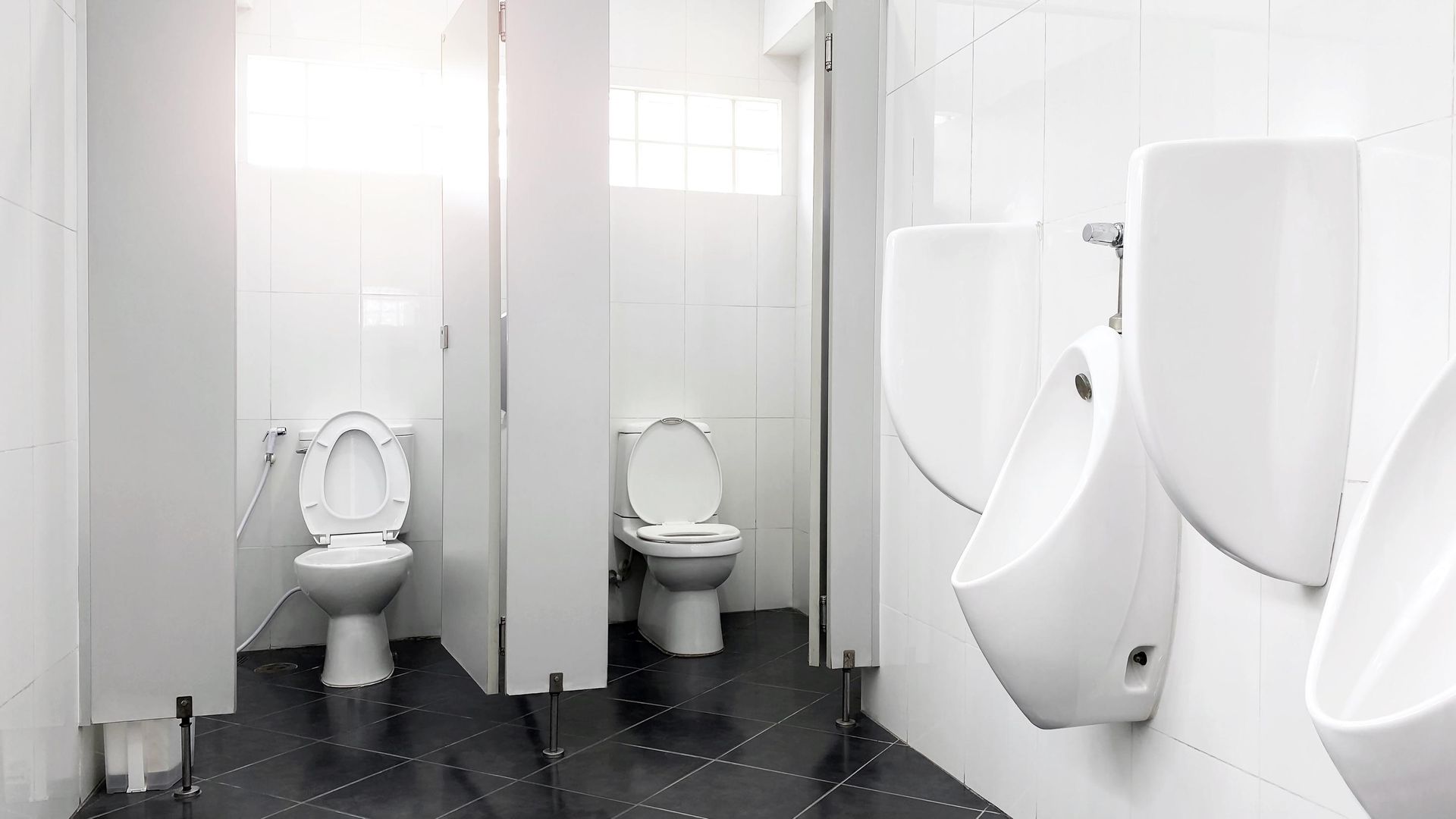 Public restroom with white urinals and toilets, separated by stalls, on a dark tiled floor.