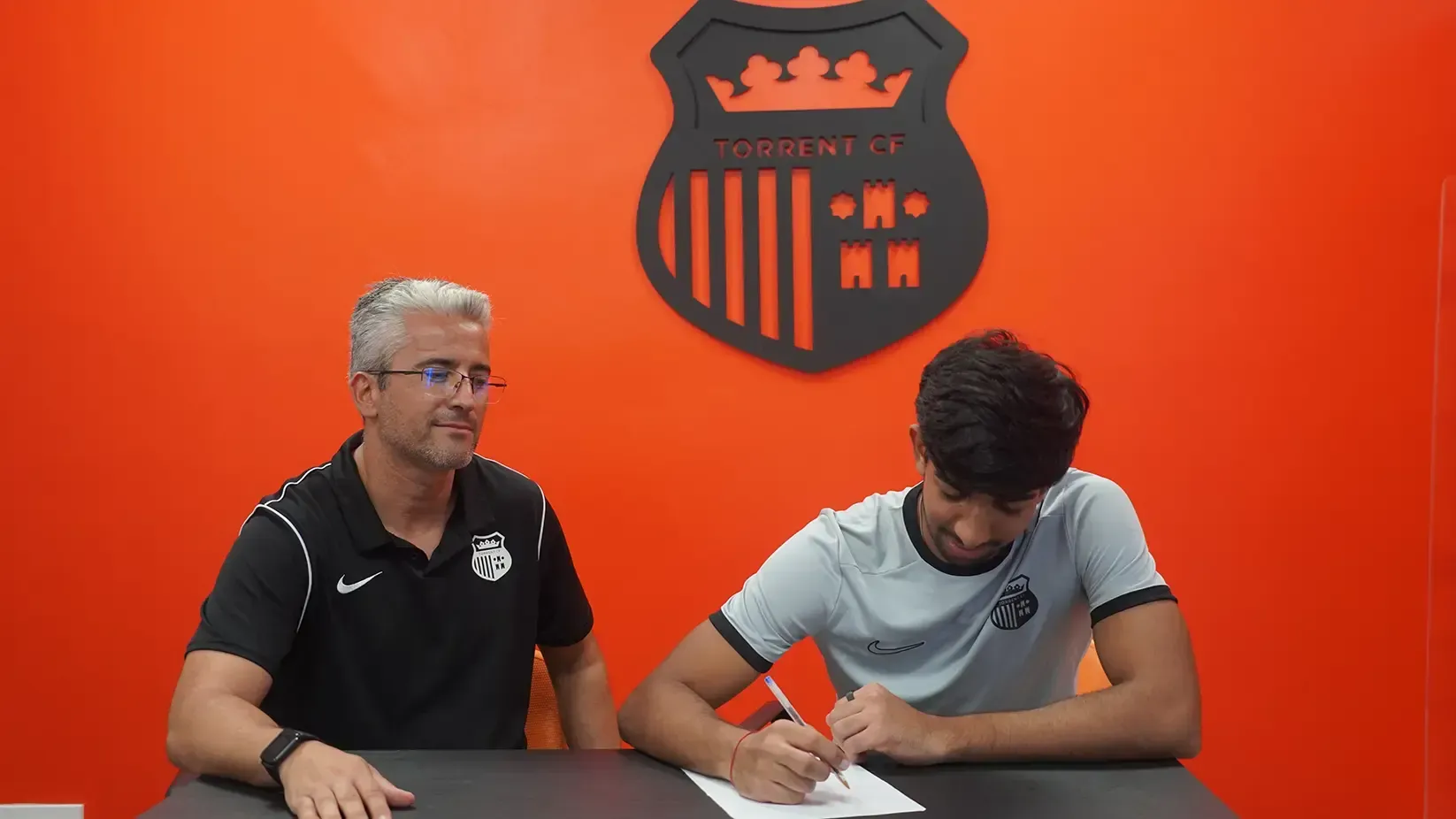 Man signs a document, seated next to another man, both in front of an orange wall with a logo.