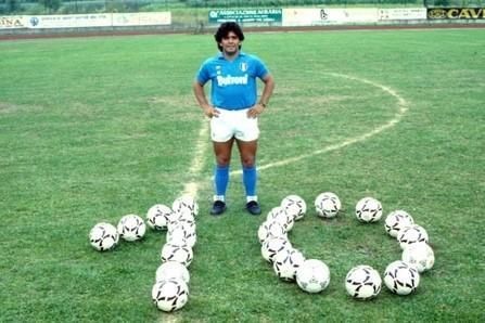 A man in a blue shirt is standing on a soccer field surrounded by soccer balls.