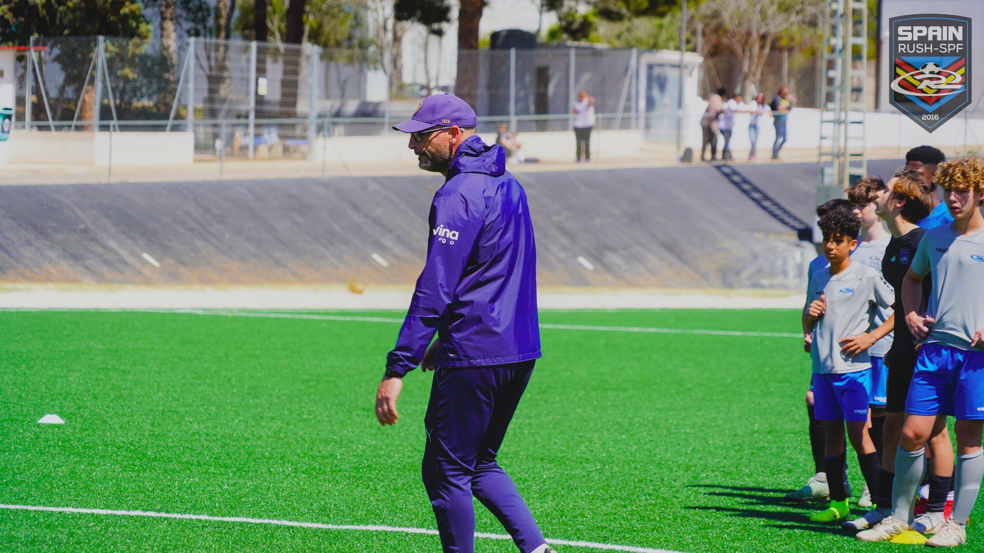 A man in a purple jacket is walking on a soccer field.