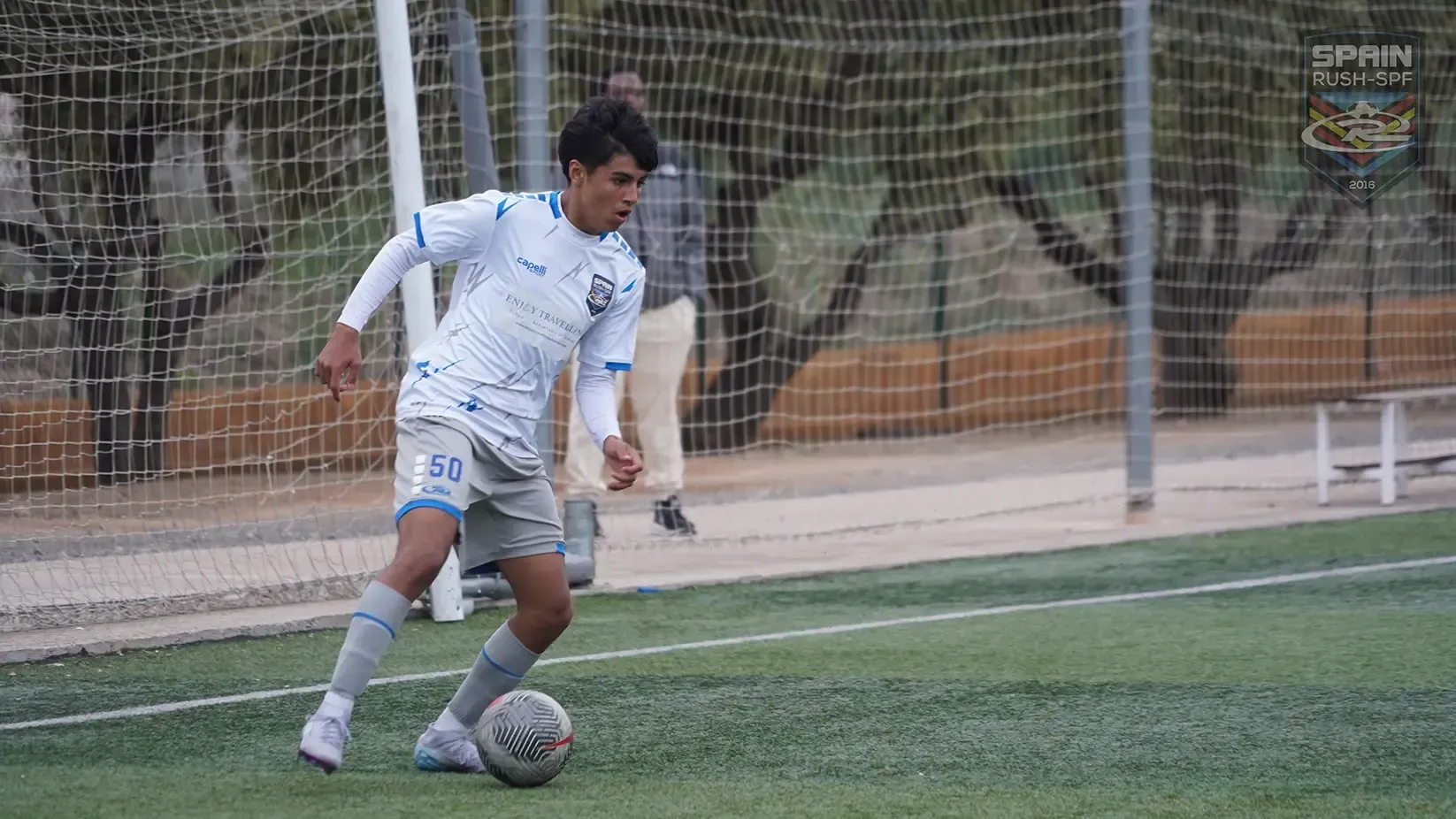 Soccer player dribbles ball on green field, wearing white and blue uniform. Net and trees in background.