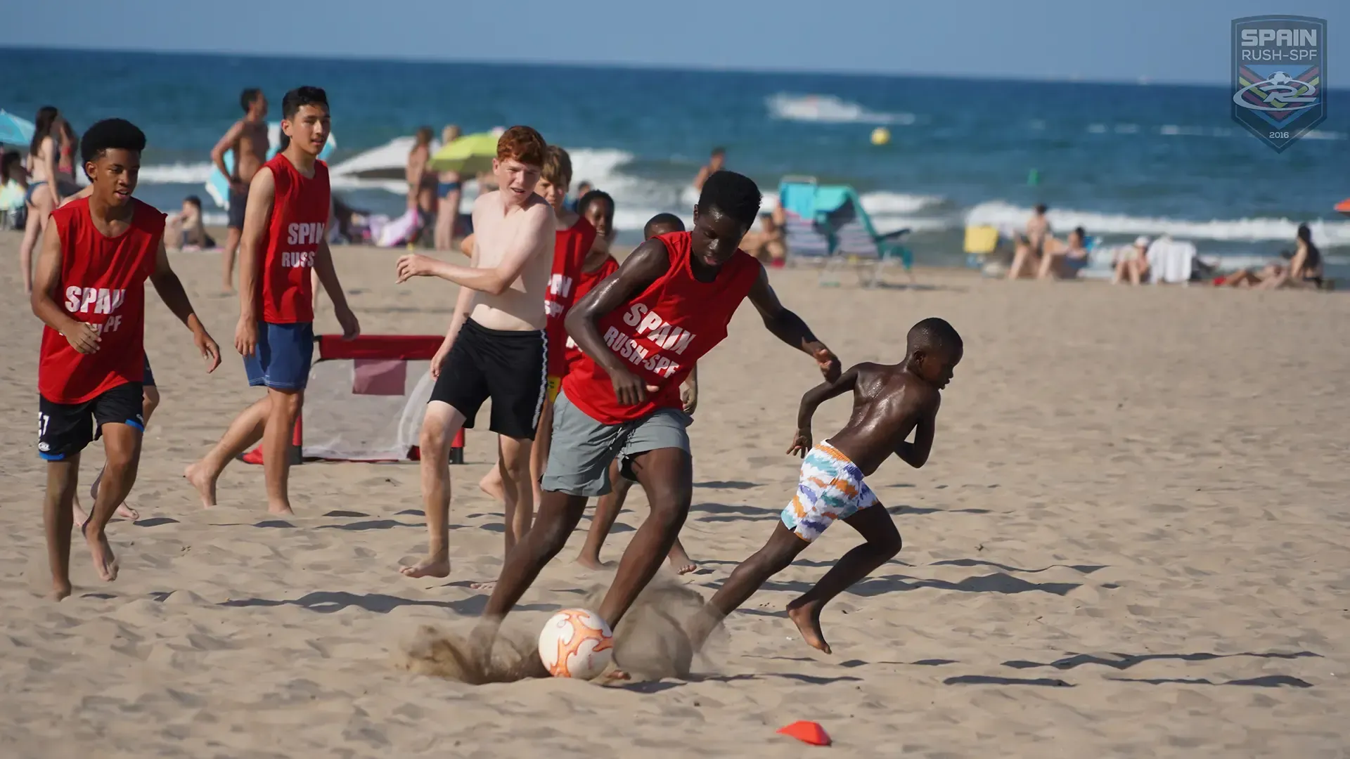 People playing soccer on a beach; one kicks a ball, others run. Sunbathers and ocean in background.