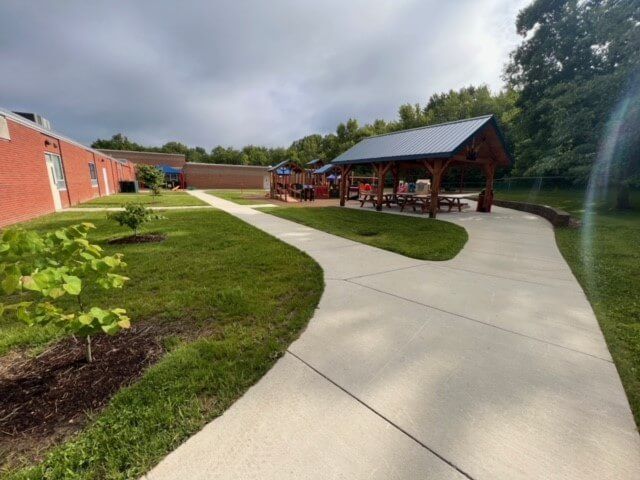 Exterior view of a schoolyard with a concrete path leading to a covered pavilion and playground.