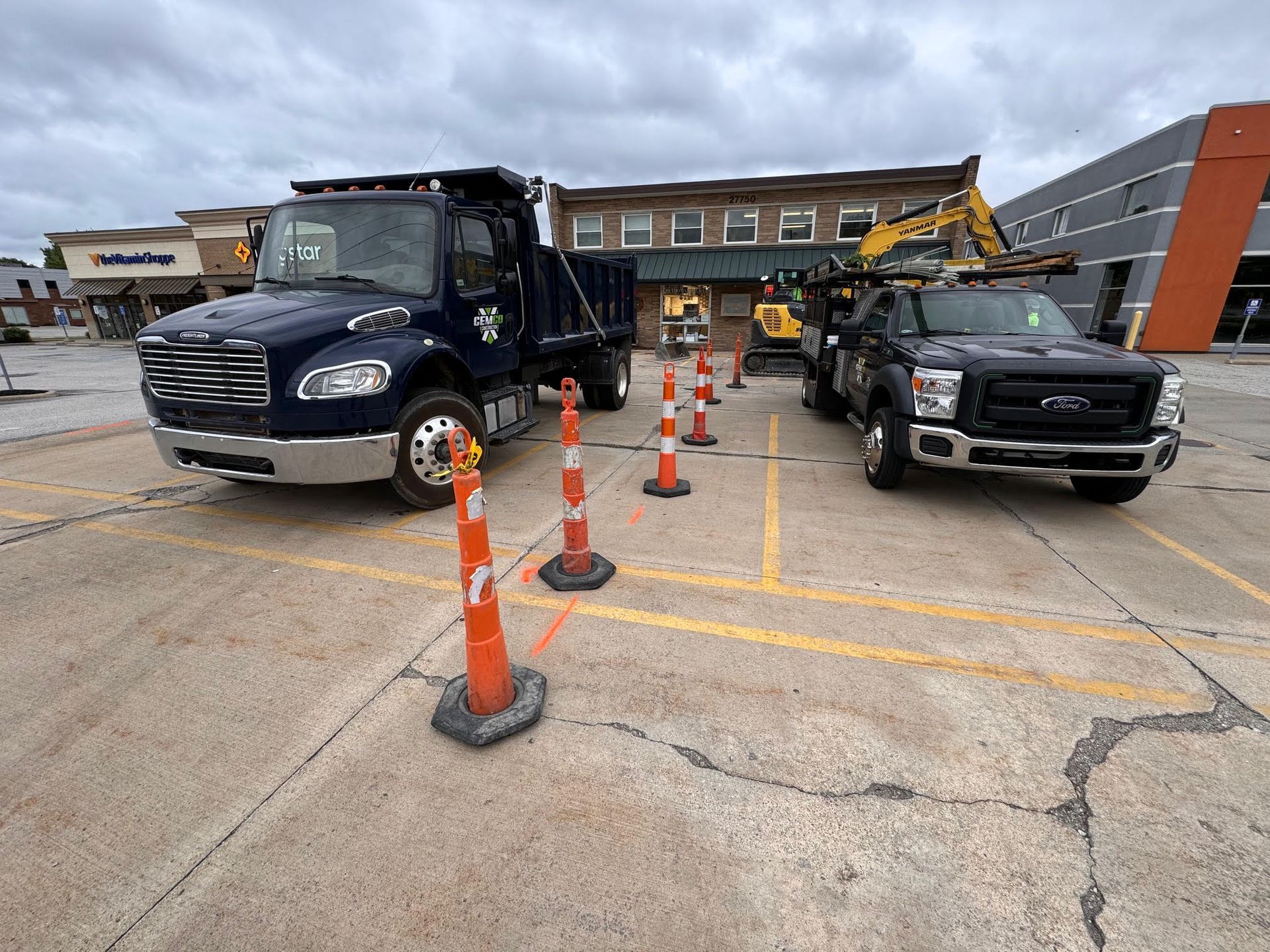 Trucks and construction equipment in a parking lot, orange cones, and a building under renovation. Cloudy day.