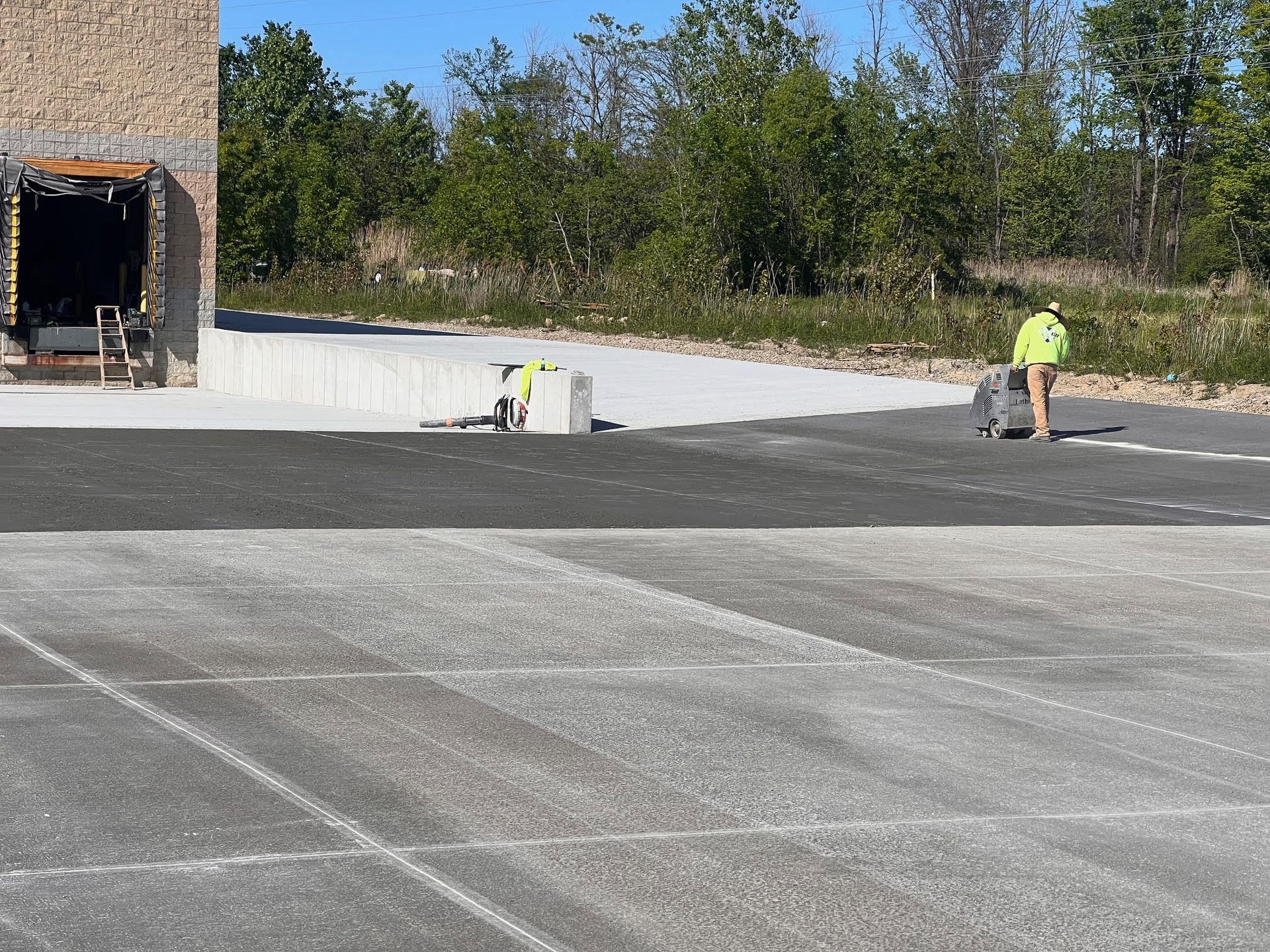 Workers using equipment on paved surface near a building and trees on a sunny day.